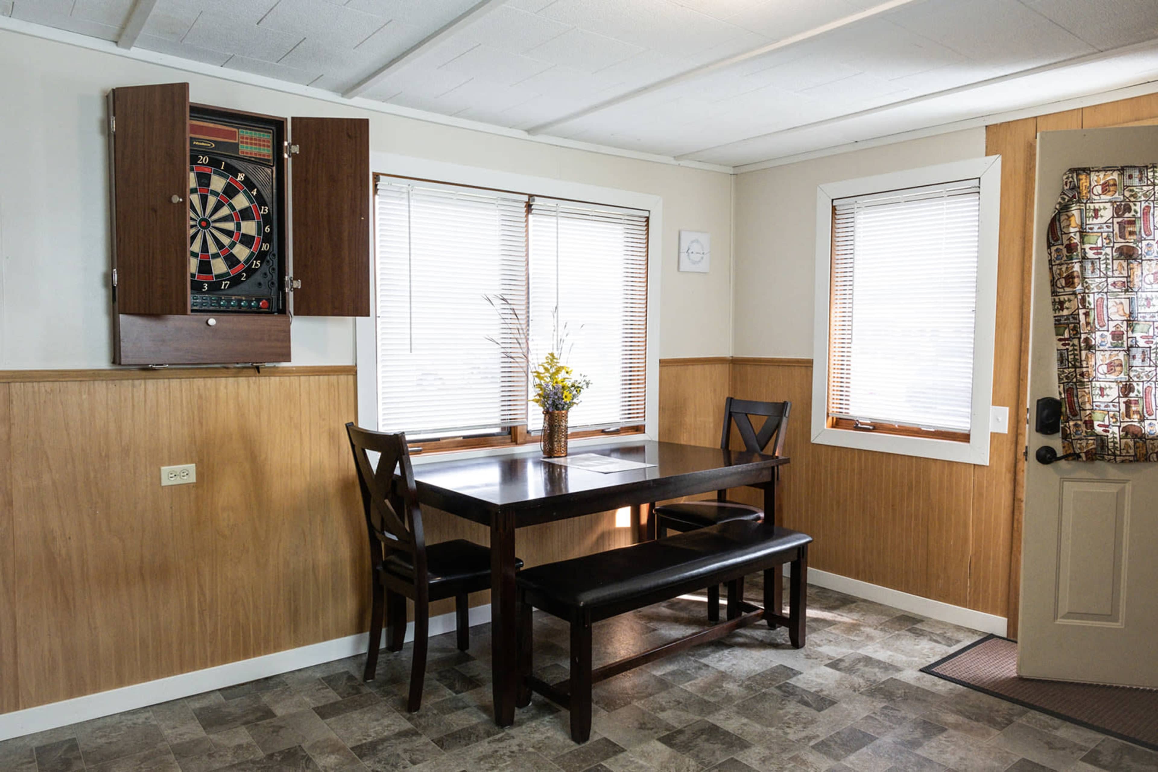 A dining area features a table with two chairs and a bench, a dartboard mounted on the wall, and a window with blinds.