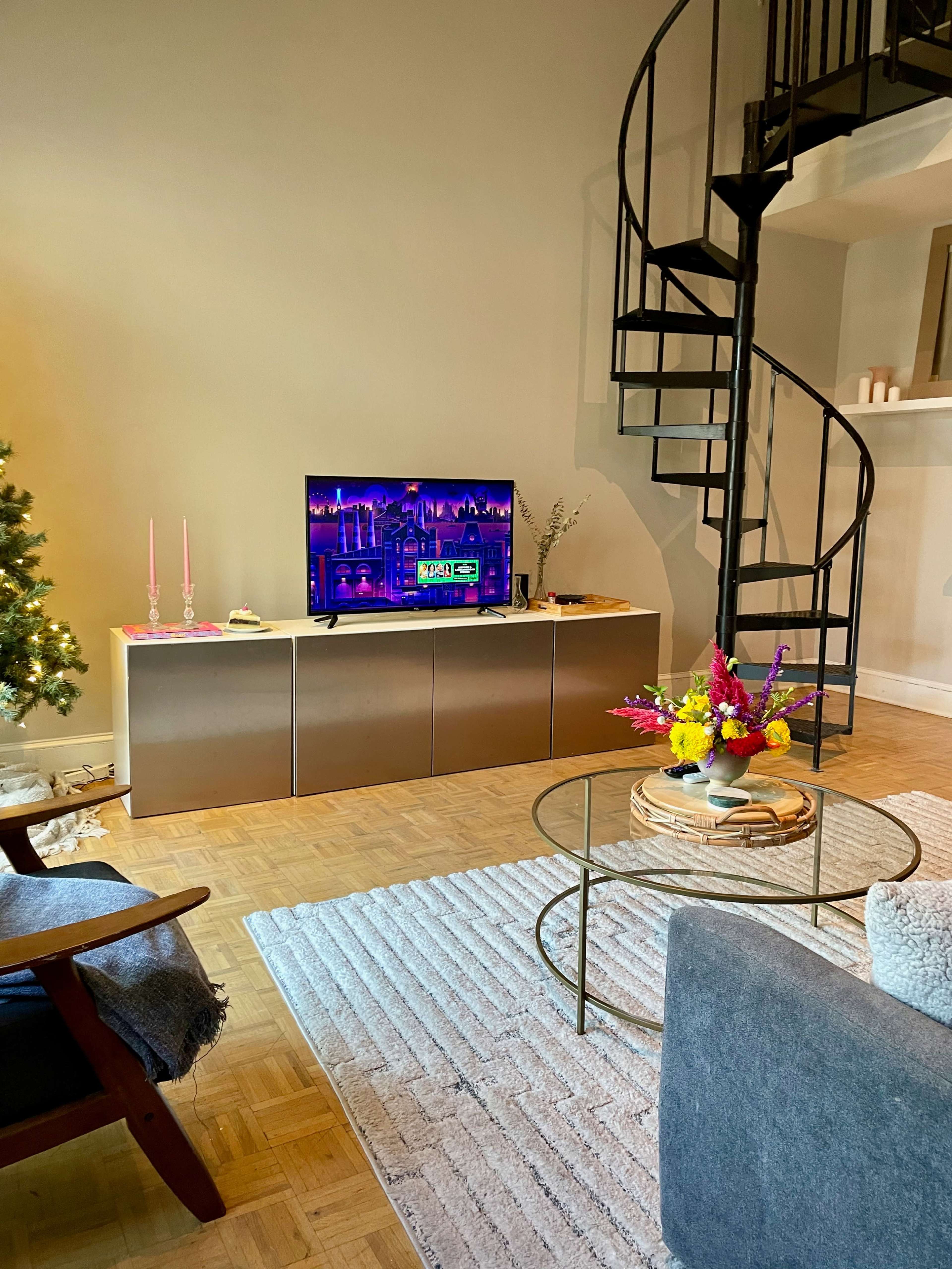 A cozy living room with a television on a cabinet, a spiral staircase in the background, a small coffee table with flowers, and a decorative rug on the wooden floor.