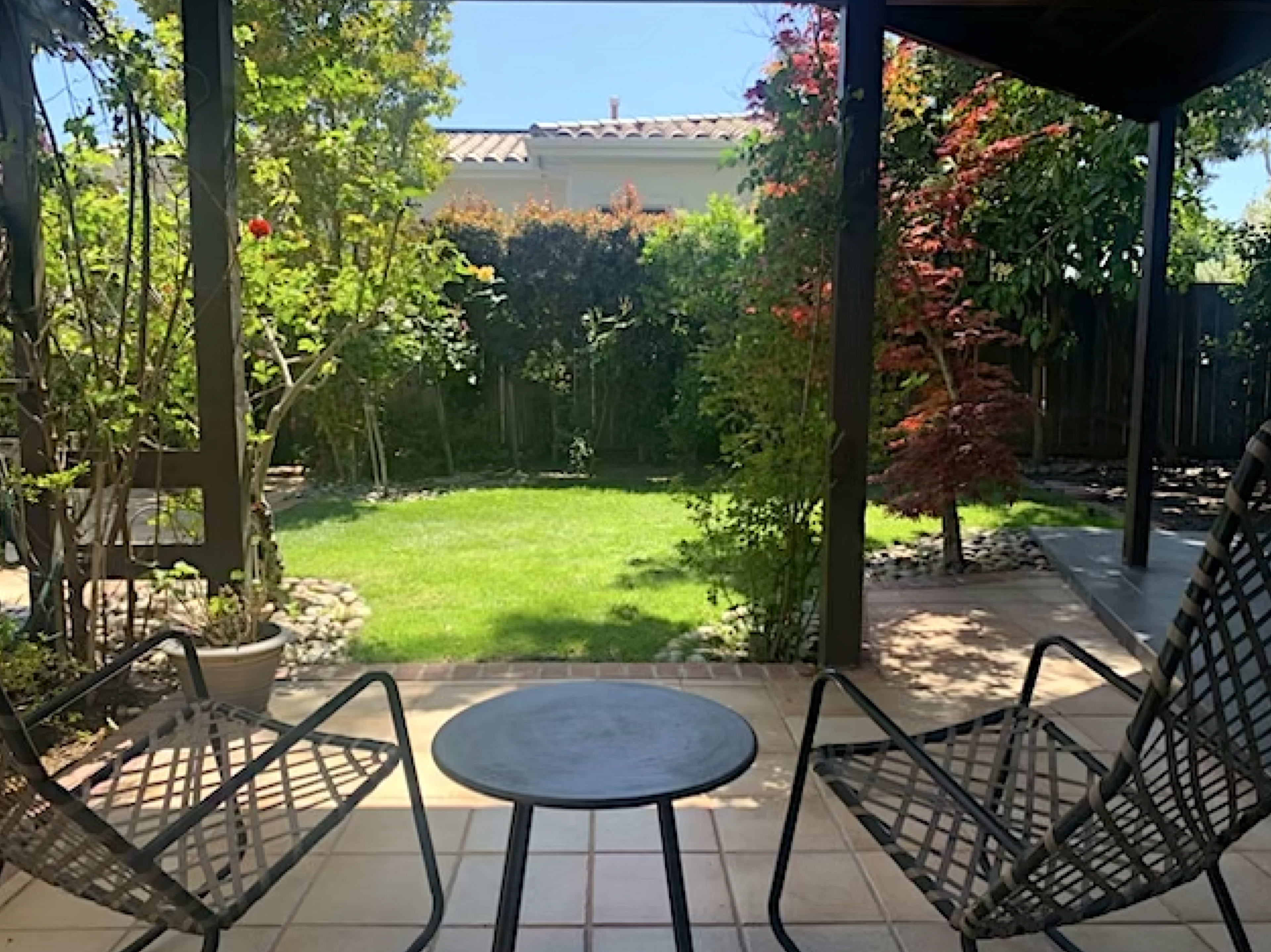 A patio with two chairs and a small table overlooking a well-maintained lawn surrounded by various plants and trees.