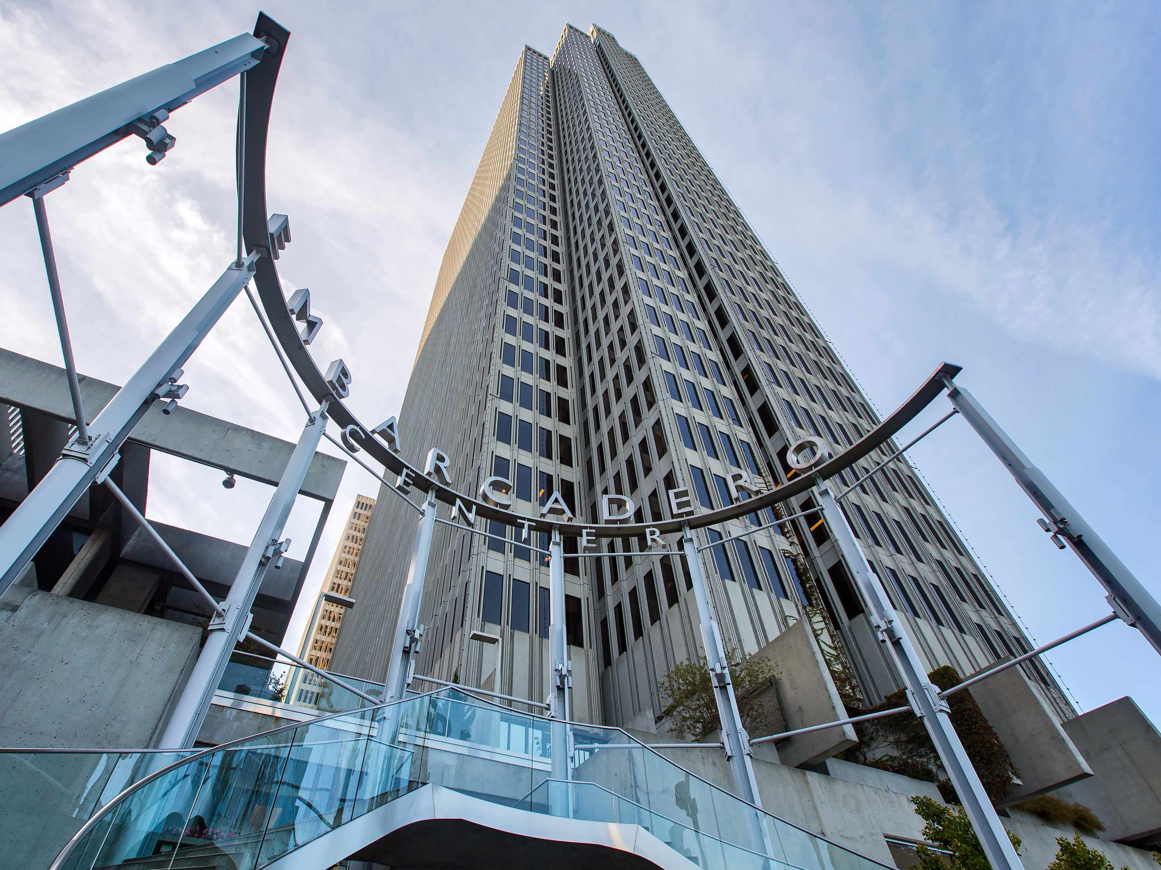 The image shows a tall skyscraper with a modern entrance structure, viewed from below, against a blue sky.
