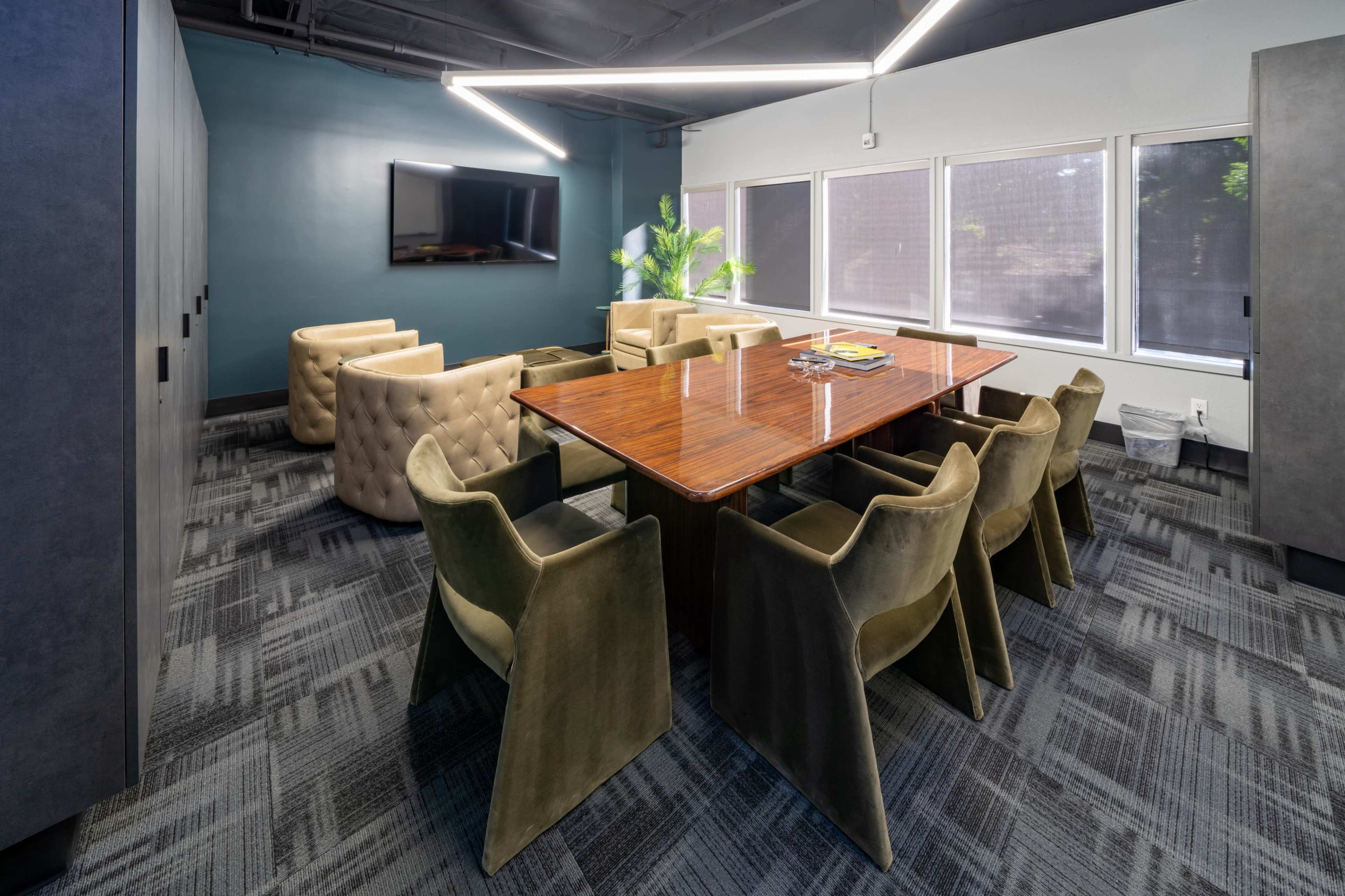 A modern conference room features a large wooden table surrounded by upholstered chairs, with a TV on the wall and large windows allowing natural light.