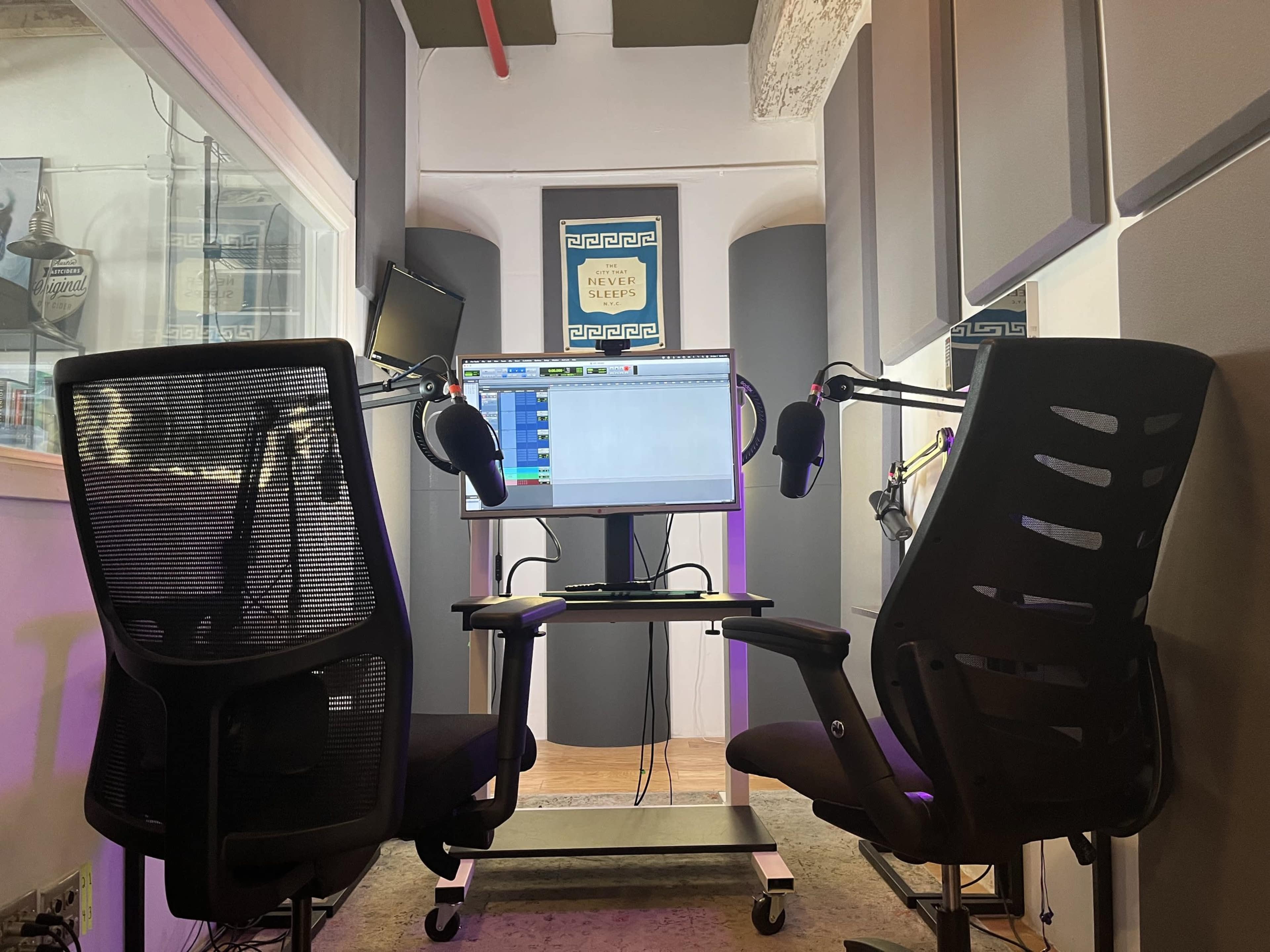 A recording studio setup features two black chairs facing a computer monitor with audio recording software, surrounded by soundproofing panels.