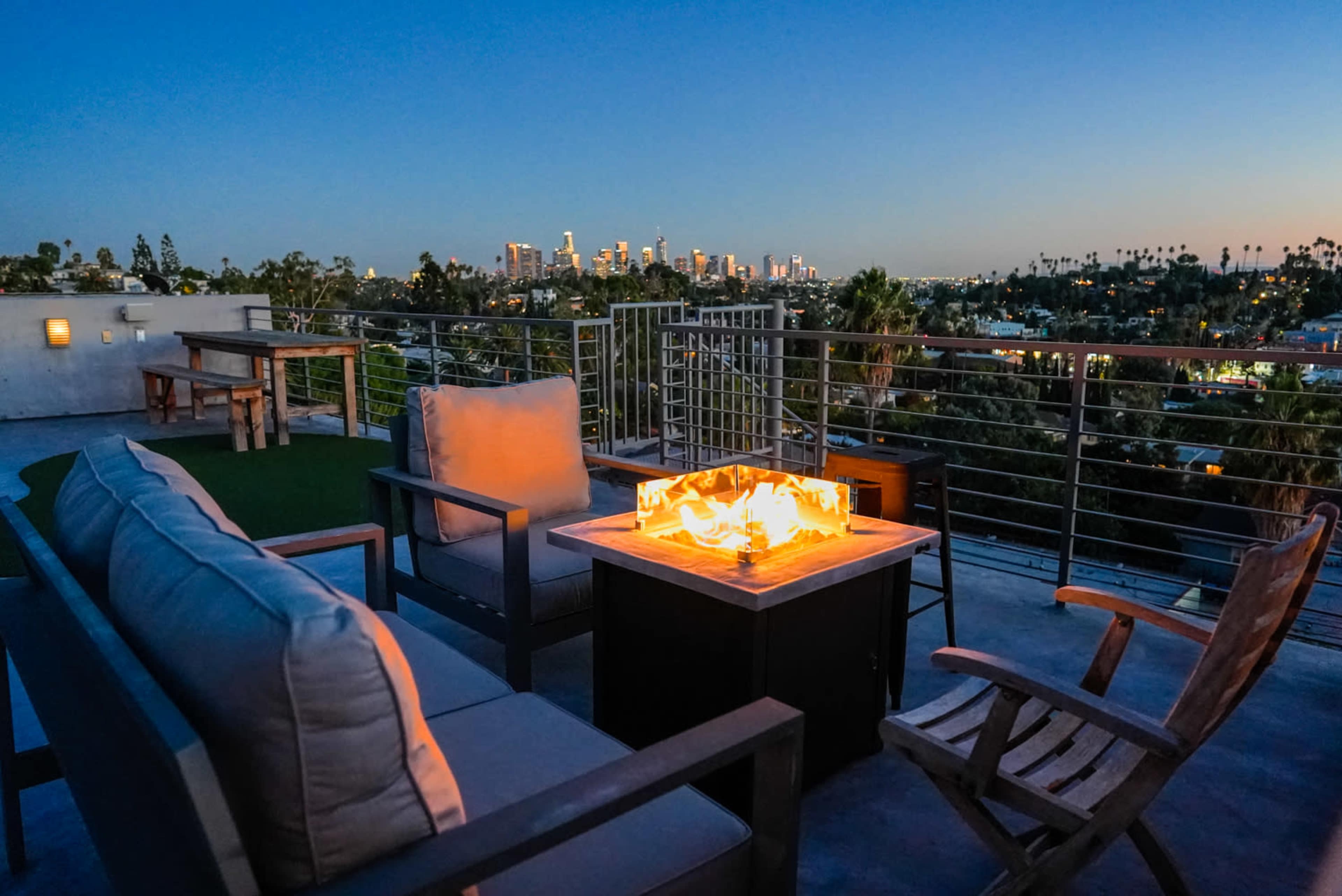 A rooftop lounge area features comfortable seating and a fire pit, with a city skyline visible in the background at dusk.