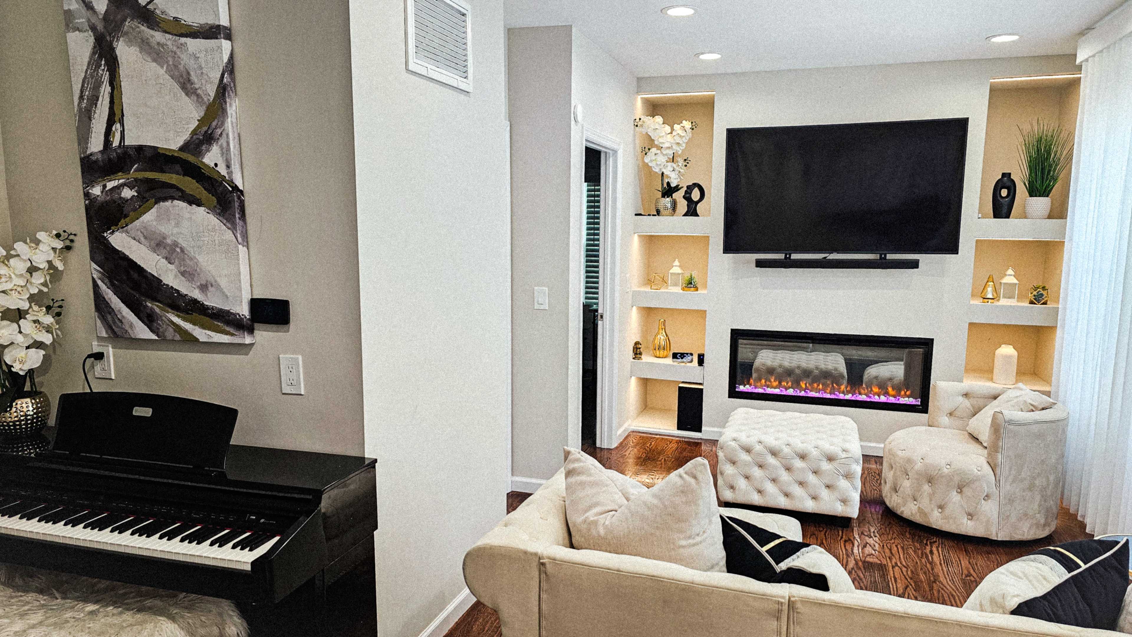 A modern living room featuring a black piano, a white tufted ottoman, a wall-mounted television above an electric fireplace, and shelves with decorative items.