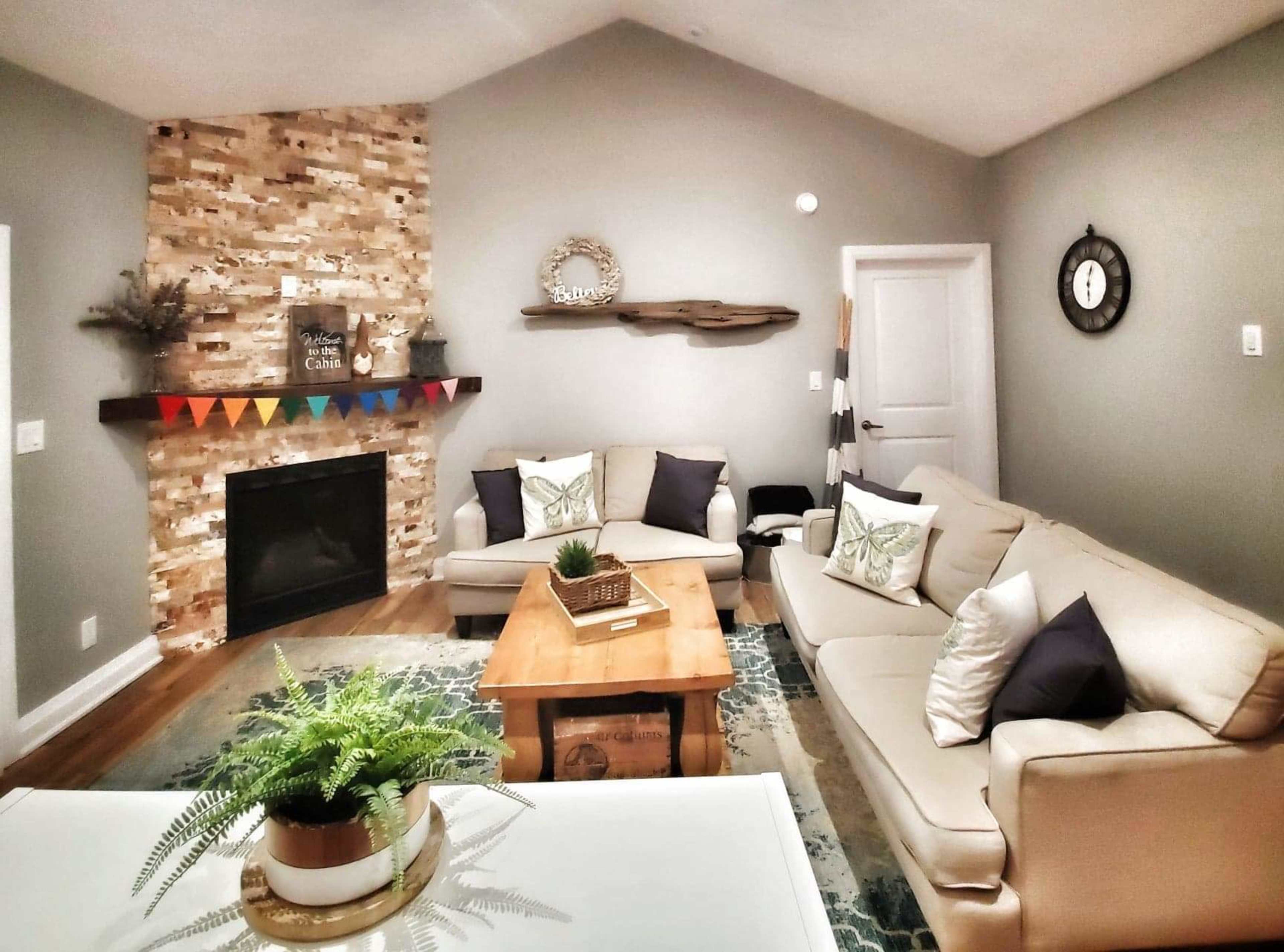 The image shows a cozy living room with a stone fireplace, beige couches, a wooden coffee table, and decorative elements on the walls.