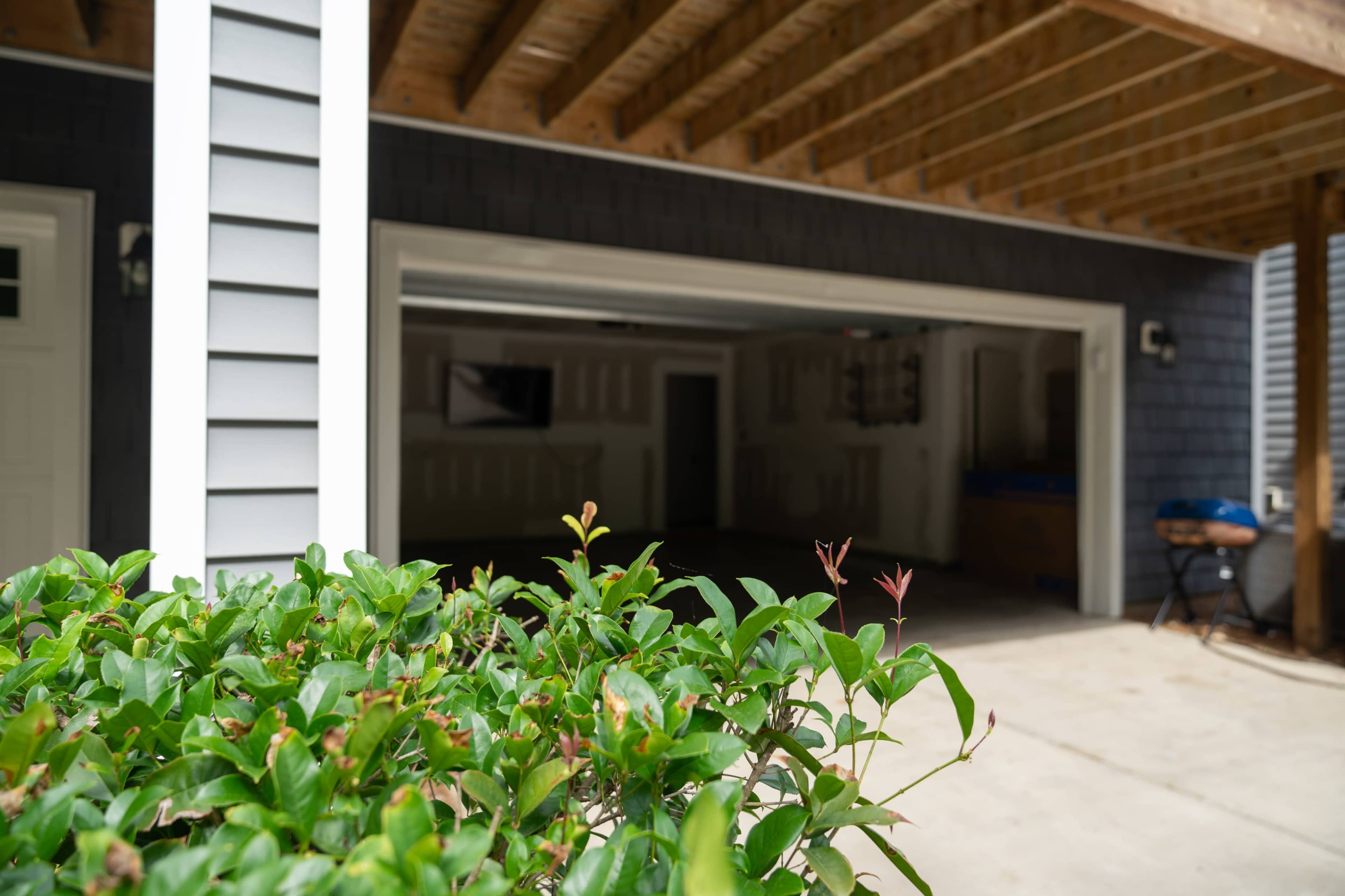 The image shows a partially enclosed garage with a view of a bush in the foreground and an interior that appears to be empty and dimly lit.