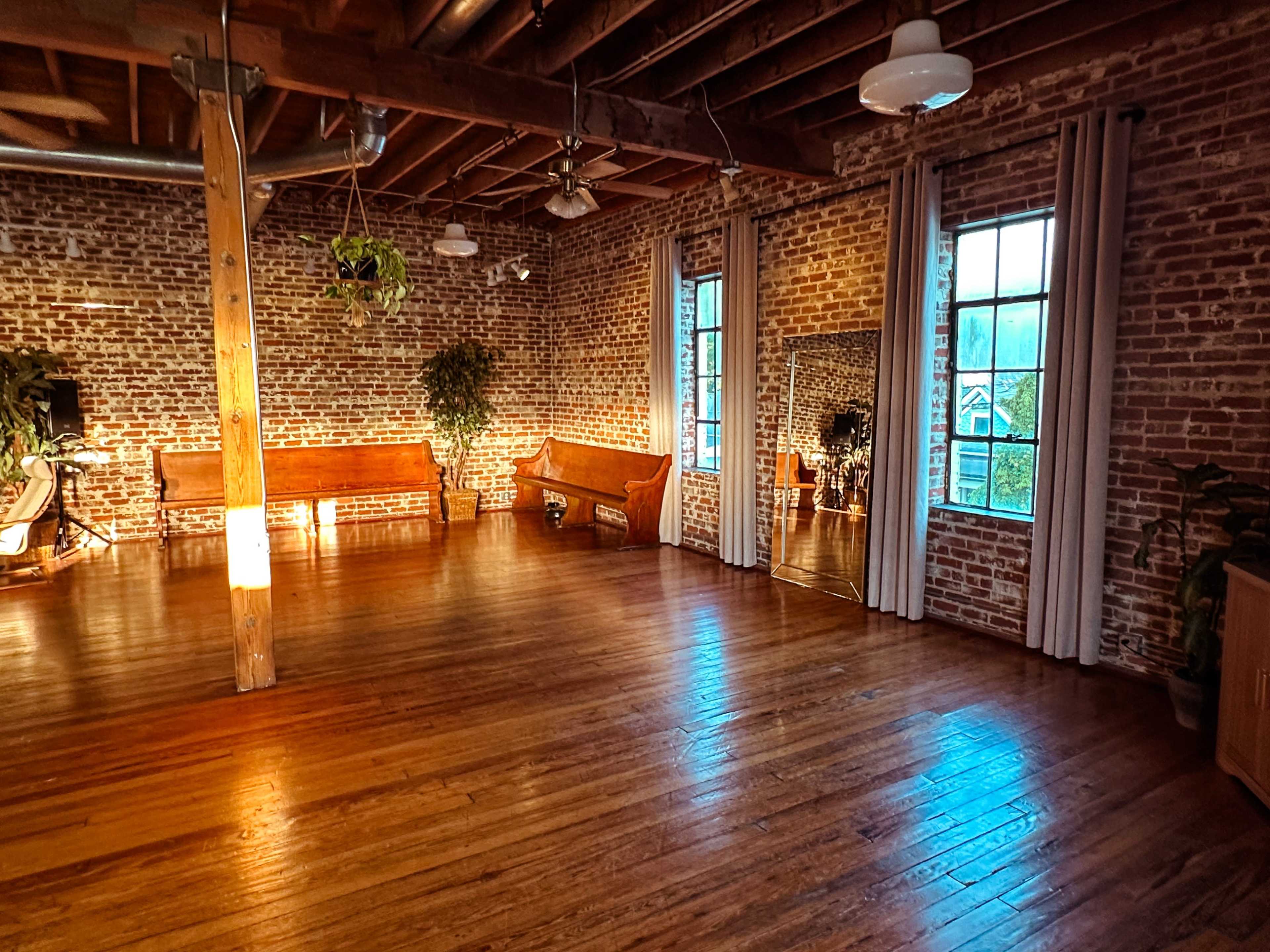 The image depicts an empty room with exposed brick walls, large windows, wooden floors, and a simple mirror reflecting some greenery.