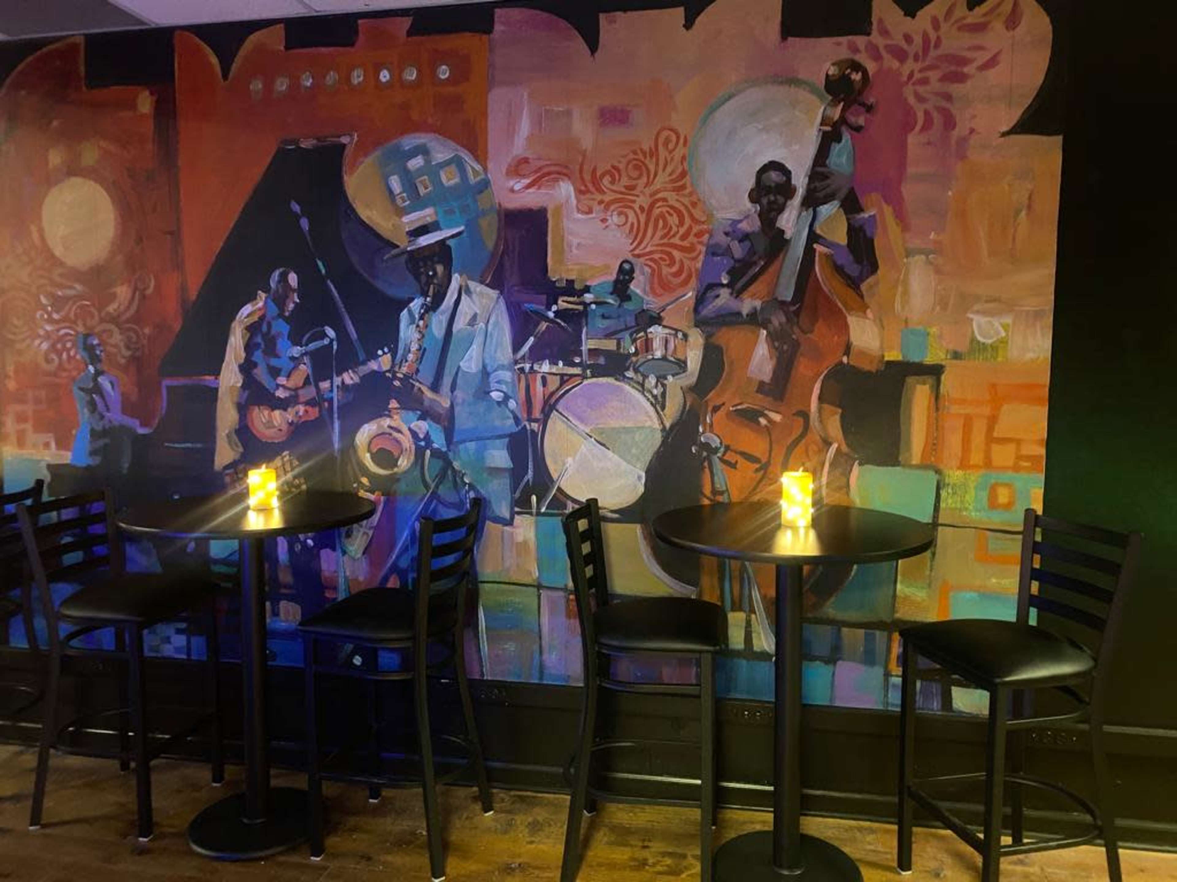 A vibrant mural of a jazz band on a wall behind empty black tables and chairs in a dimly lit venue.