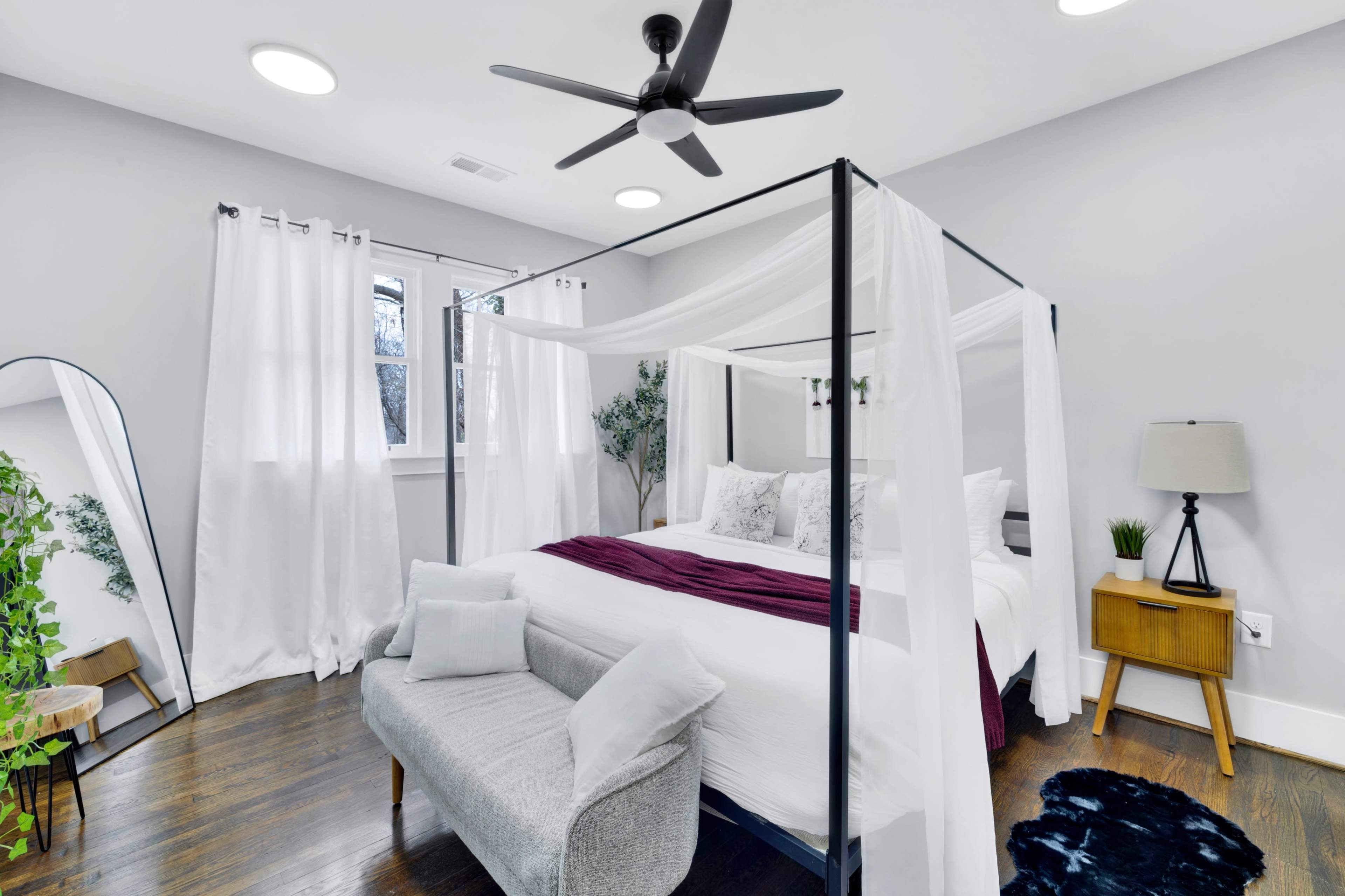 A modern bedroom features a four-poster bed with sheer drapery, a gray sofa, and a mix of decorative elements including plants and a large mirror.