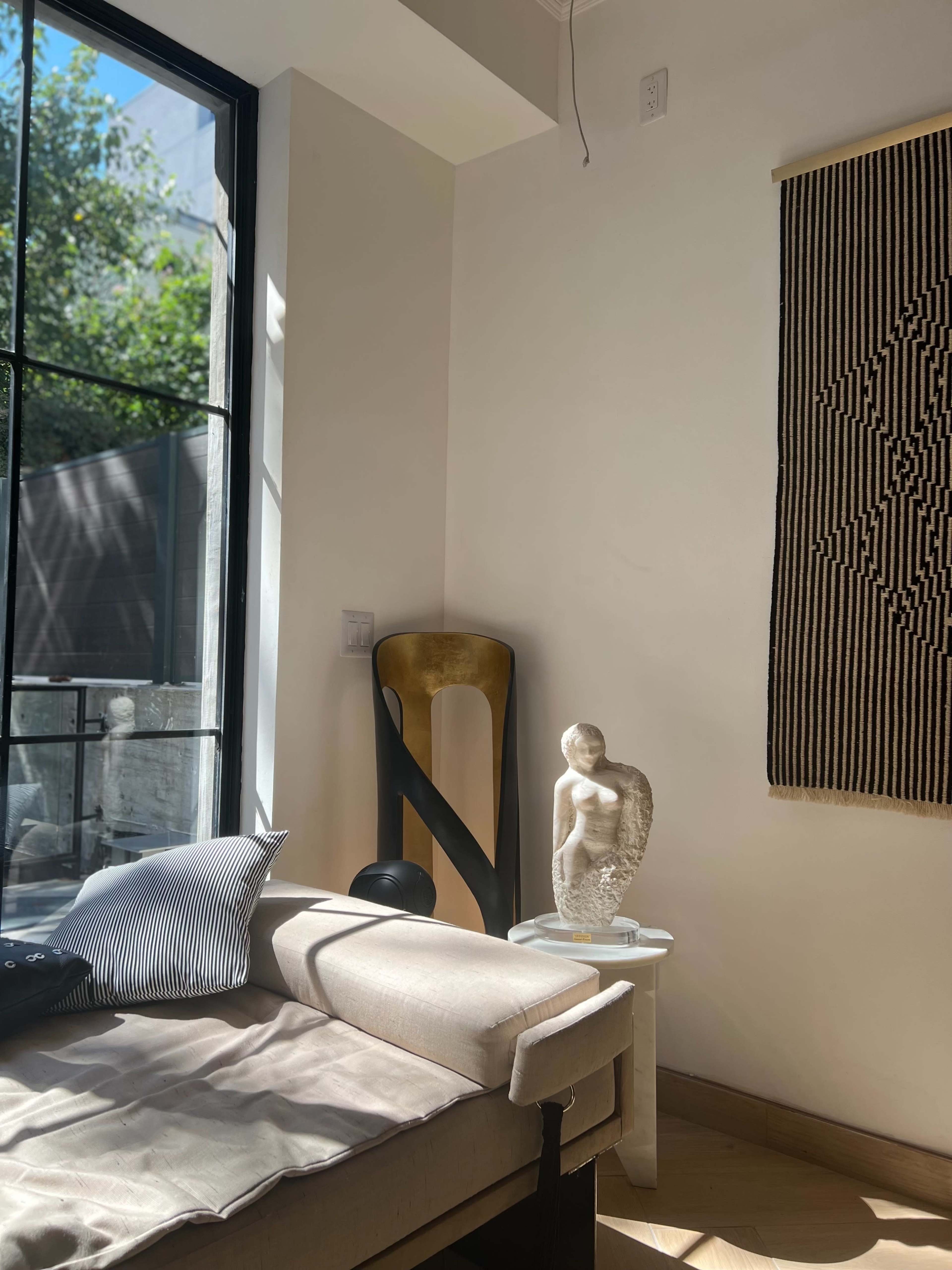 The image shows a sunlit interior featuring a modern couch with cushions, a decorative sculpture on a pedestal, and a wall hanging.