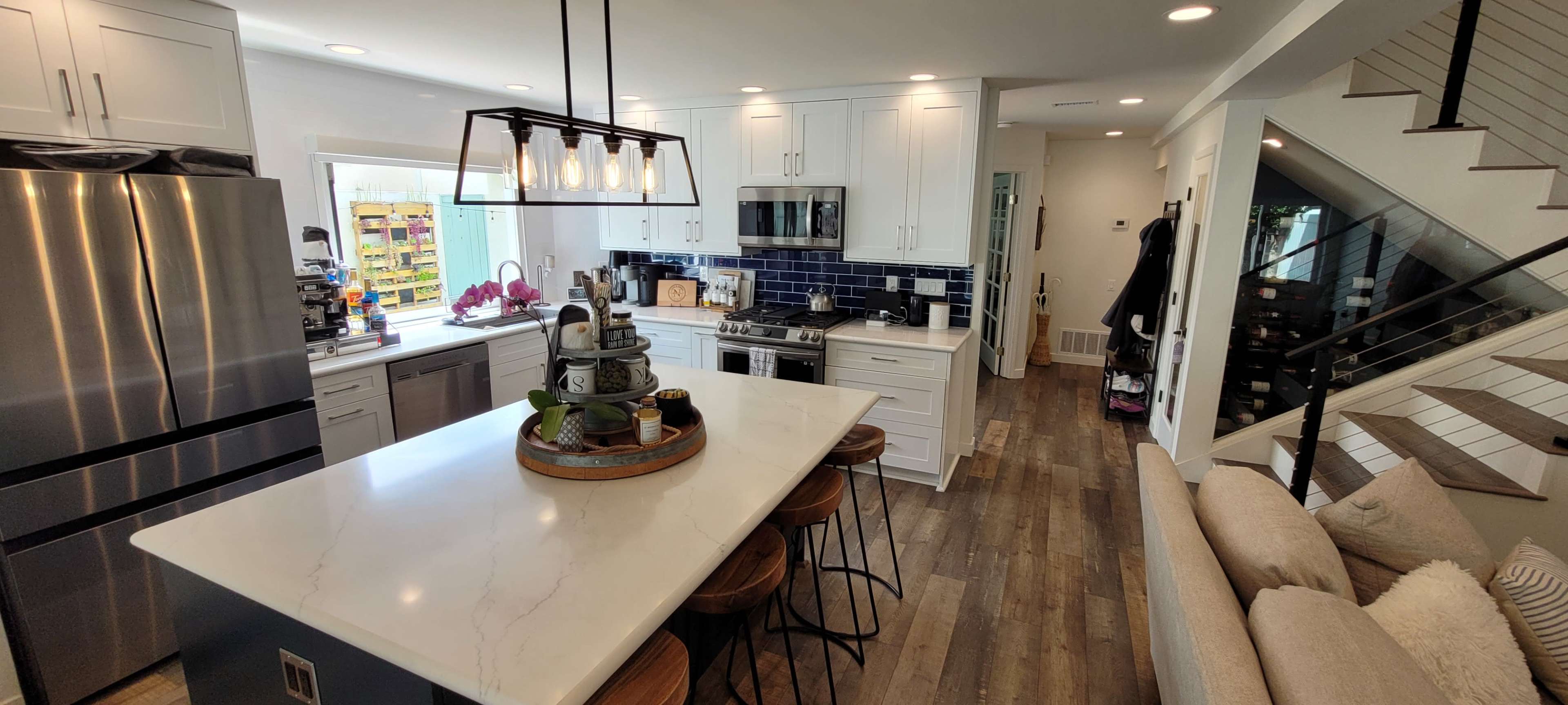 A modern kitchen with a central island, surrounded by stainless steel appliances and a living area featuring a comfortable sofa and staircase.