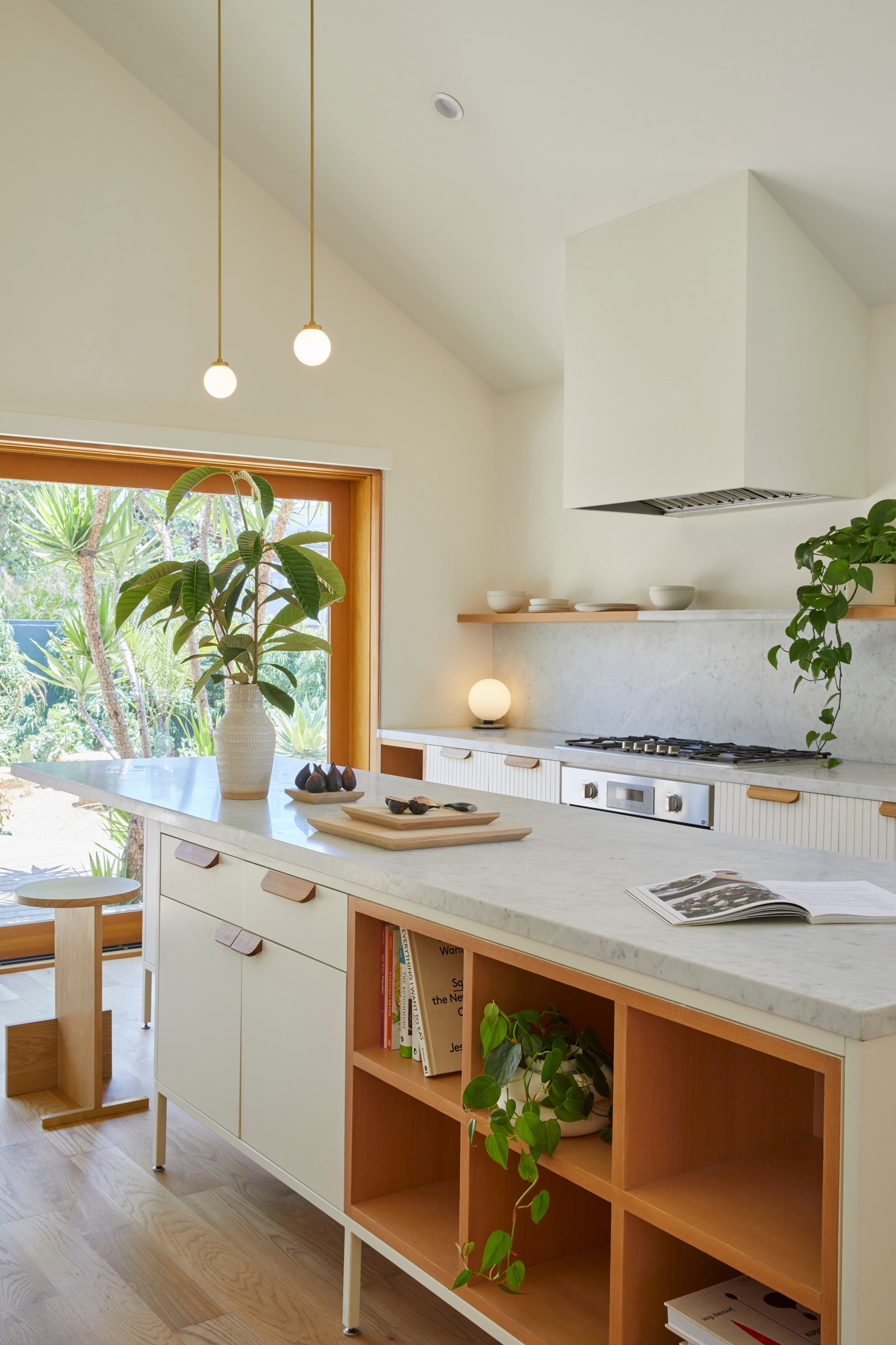 A modern kitchen features a marble countertop, wooden shelves, and plants beside a large window.