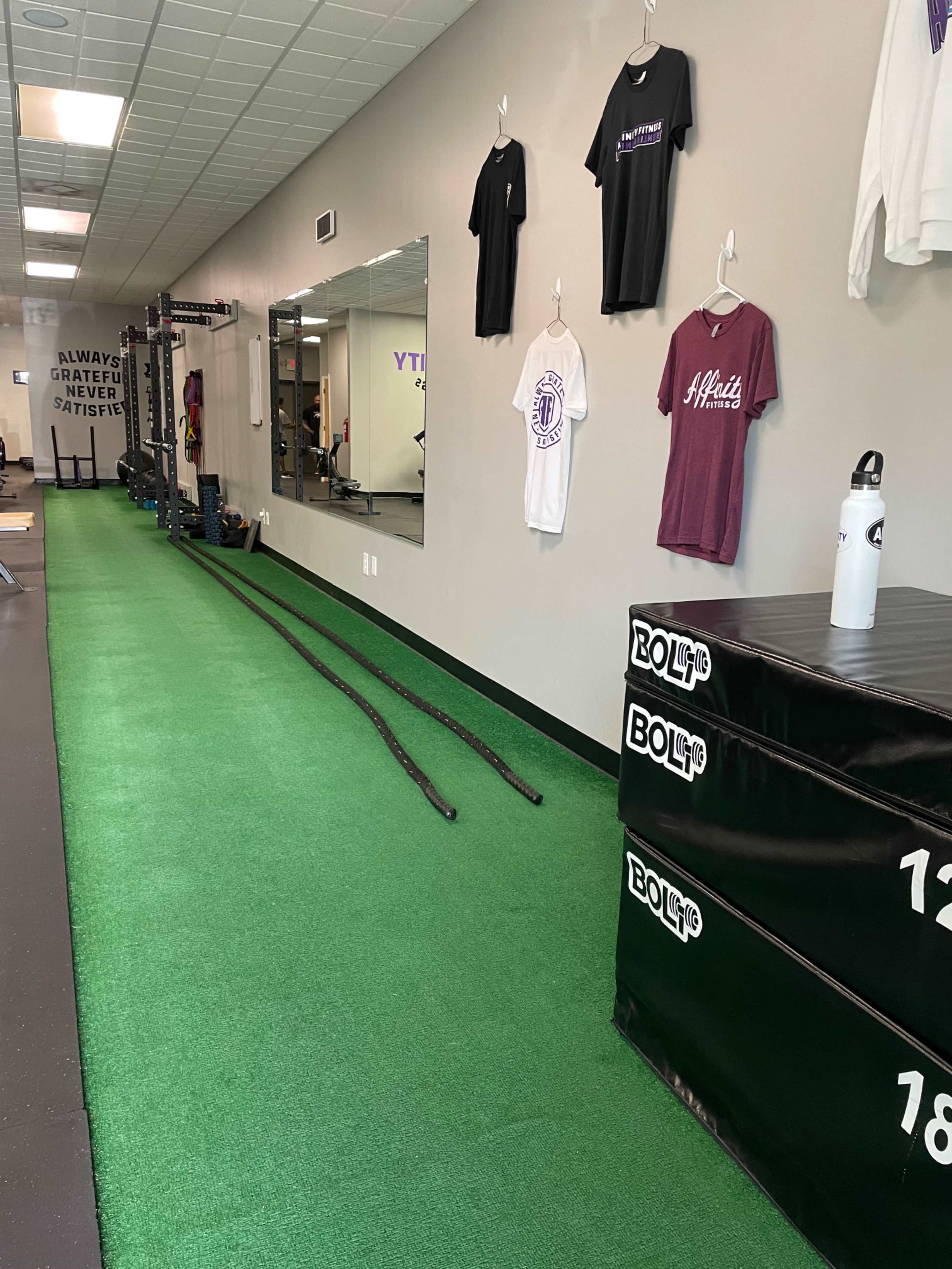 A gym interior with green turf, stacked weight boxes, exercise ropes, and workout gear hanging on the walls.