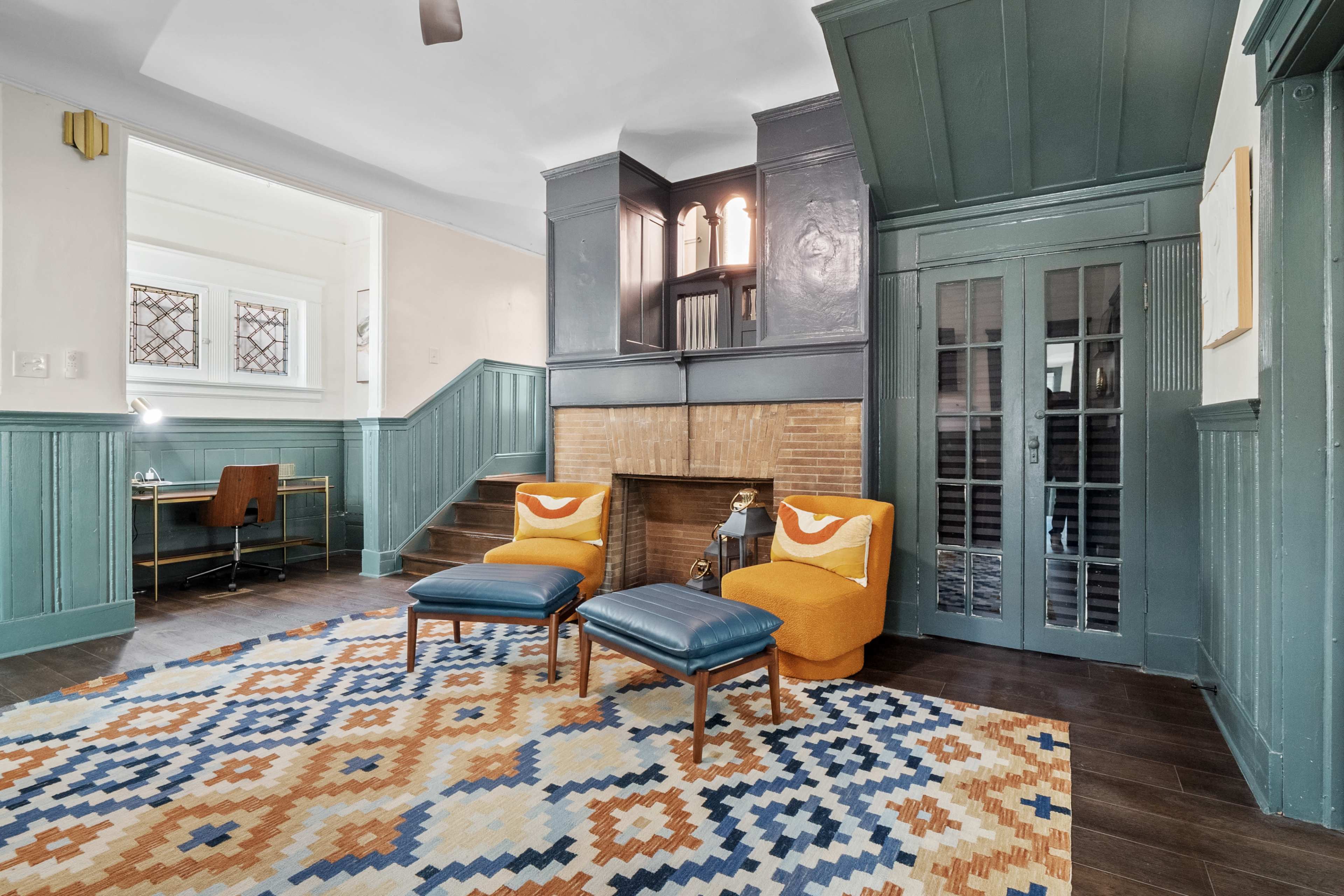 A living room with a patterned rug, two orange and blue chairs, and a fireplace with decorative panels.