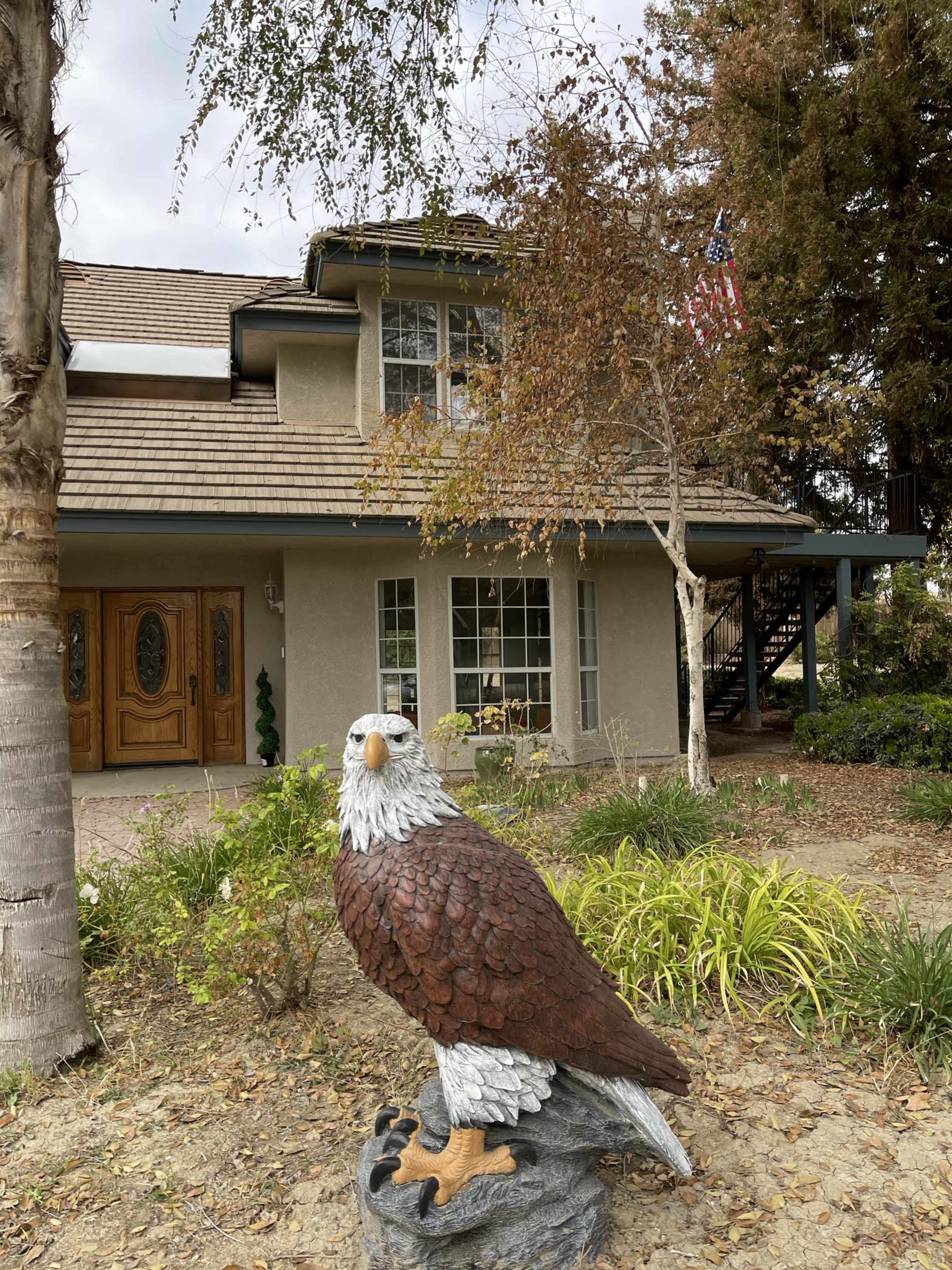 A large eagle statue stands in front of a two-story house with a staircase and an American flag hanging from the porch.
