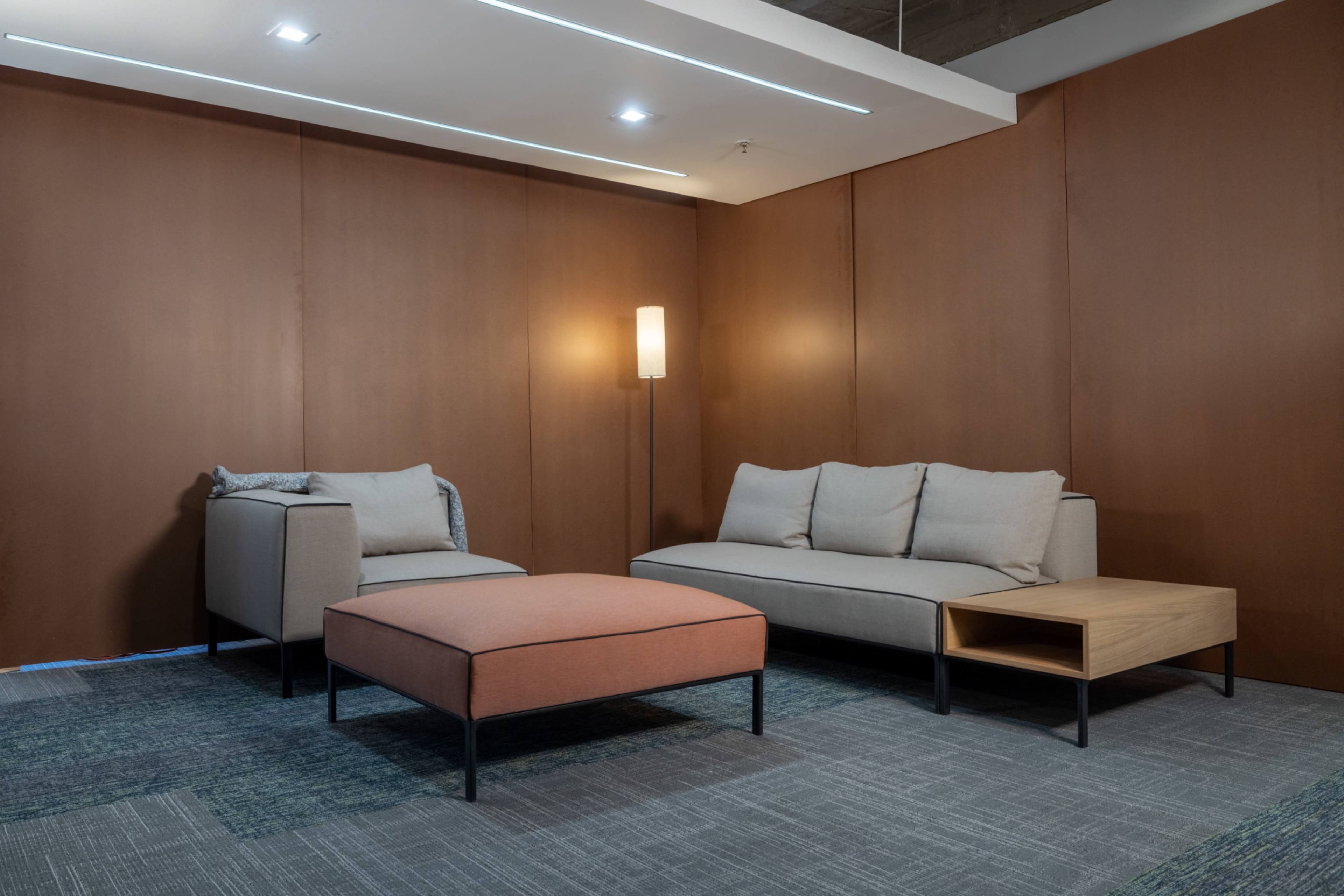 The image features a modern living room setup with a beige sofa, an armchair, a light wooden side table, an orange ottoman, and a standing lamp against a brown paneled wall.