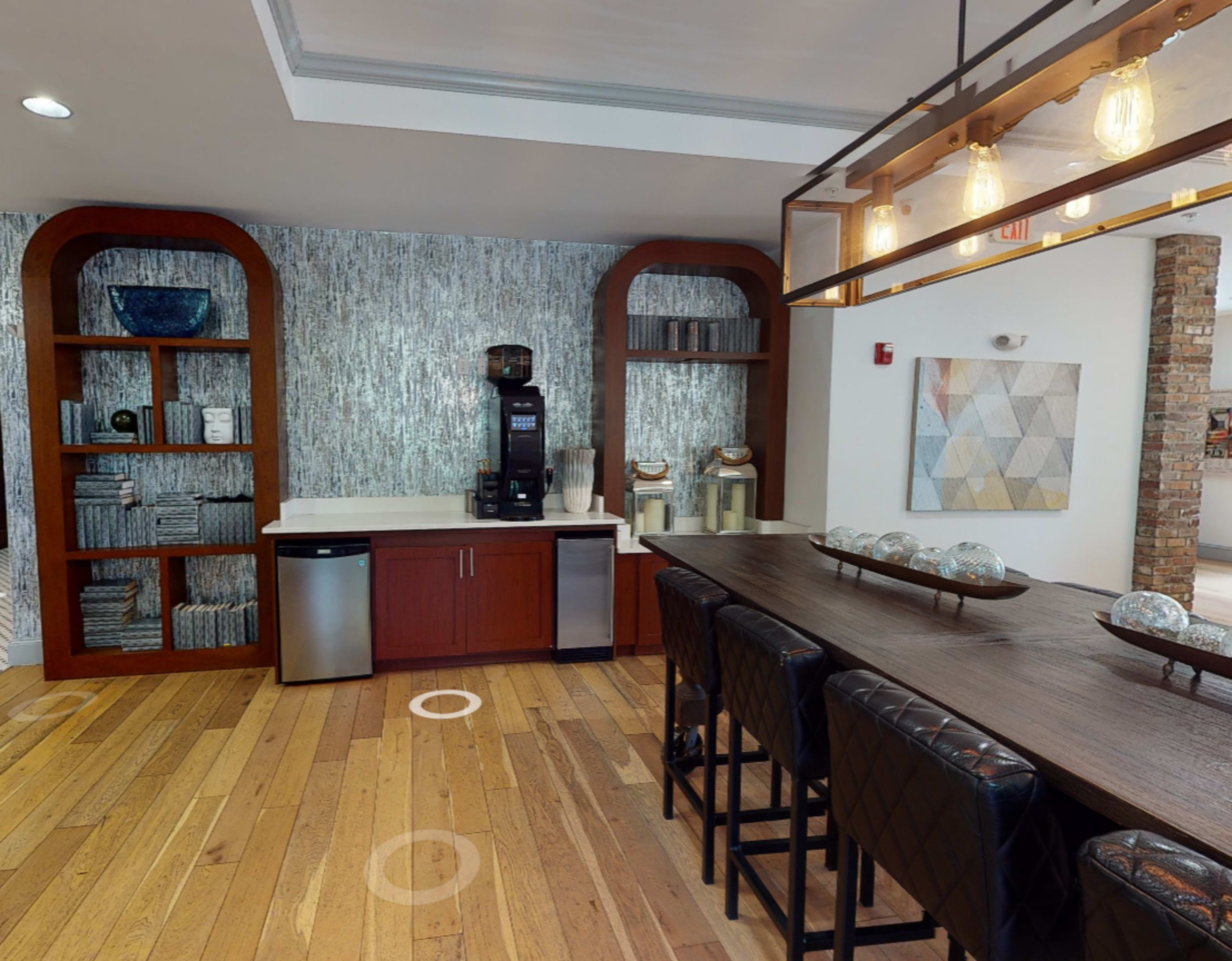 A modern kitchen area with wooden flooring, featuring a coffee machine on a countertop, shelves filled with towels, and a long dining table with black chairs.