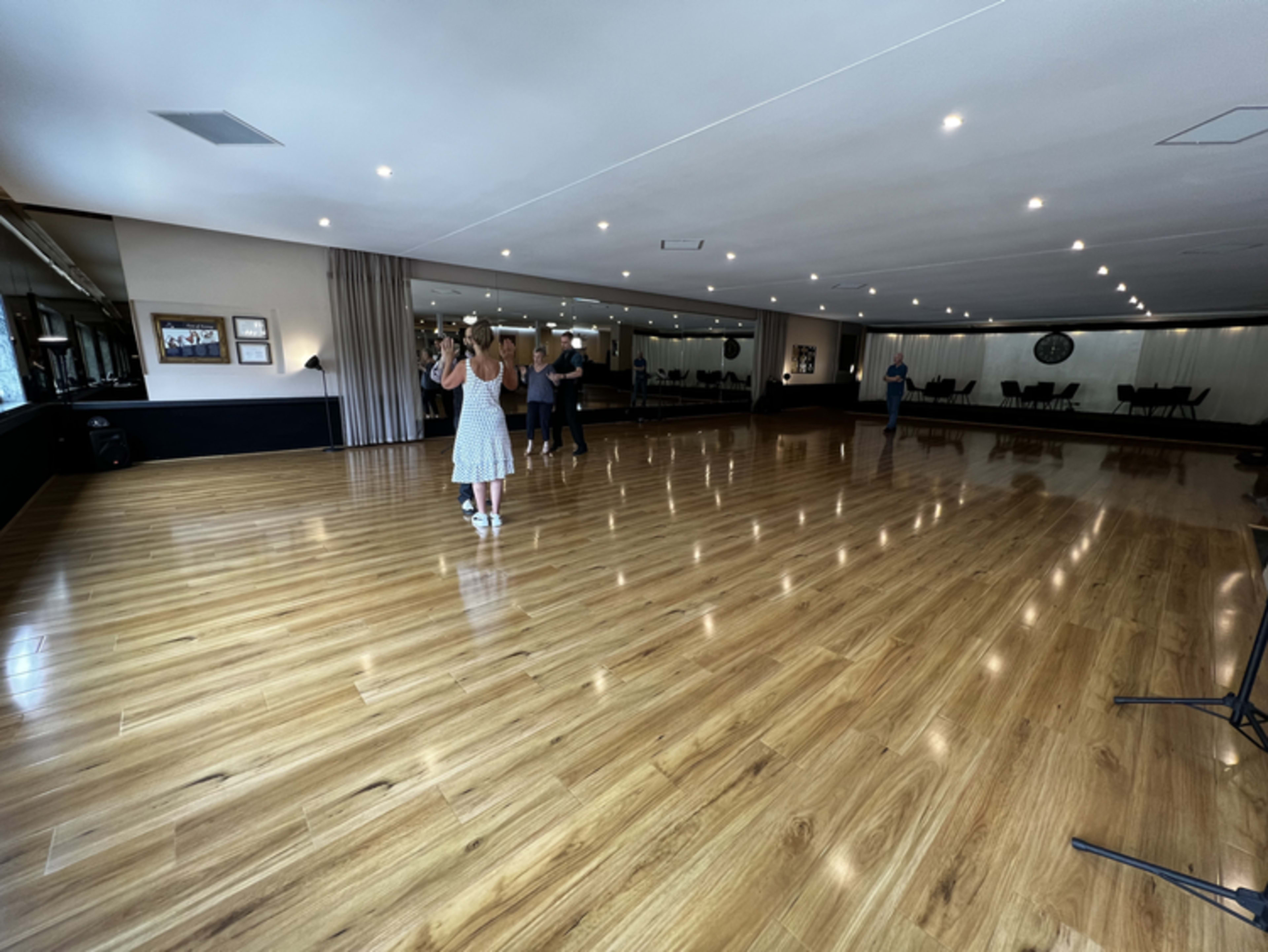 A spacious dance studio with polished wooden floors, mirrors on one wall, and attendees practicing in pairs.