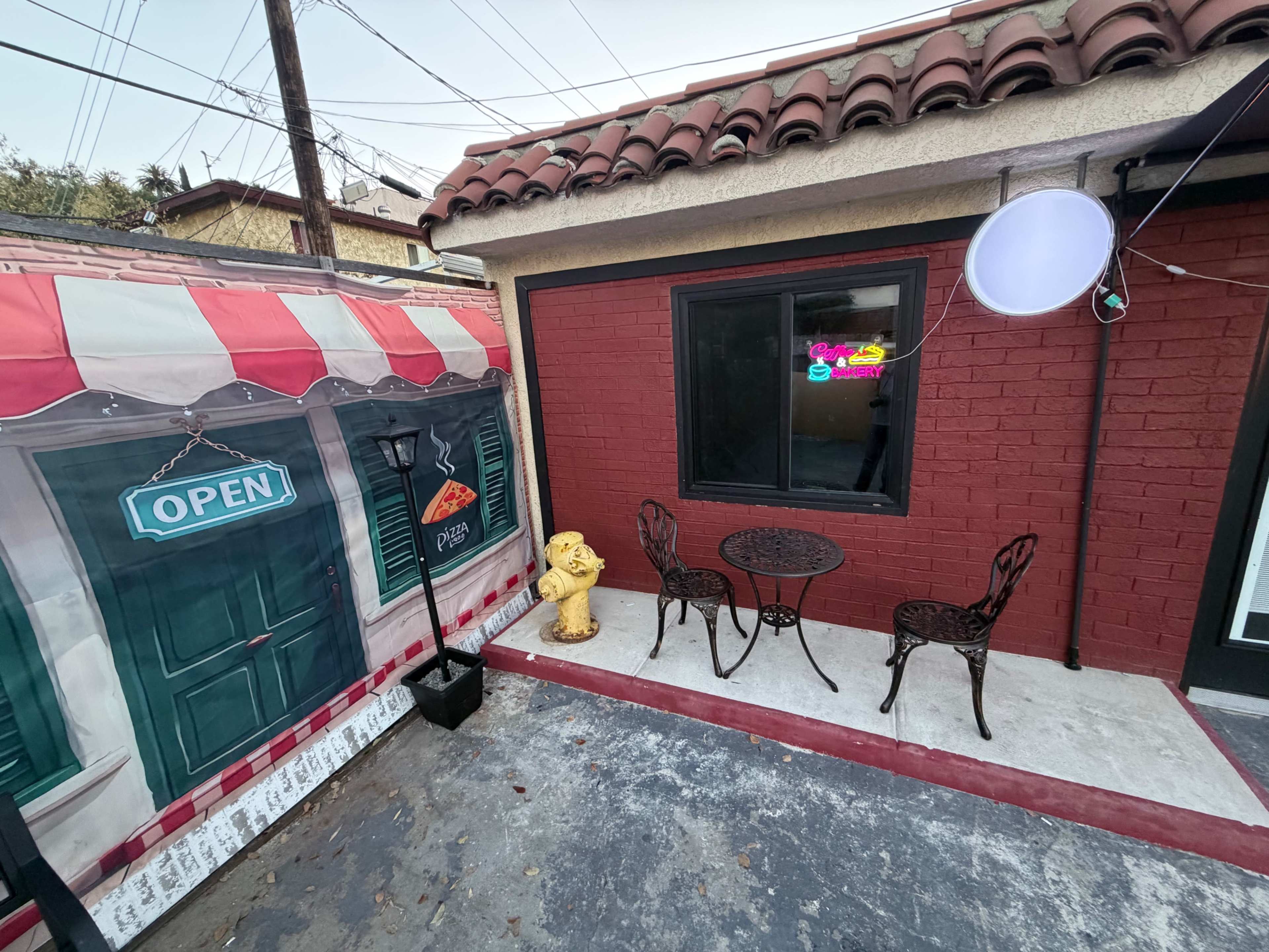 A small outdoor seating area features a round table with two wrought iron chairs alongside a decorative statue and a mural depicting a pizza shop.