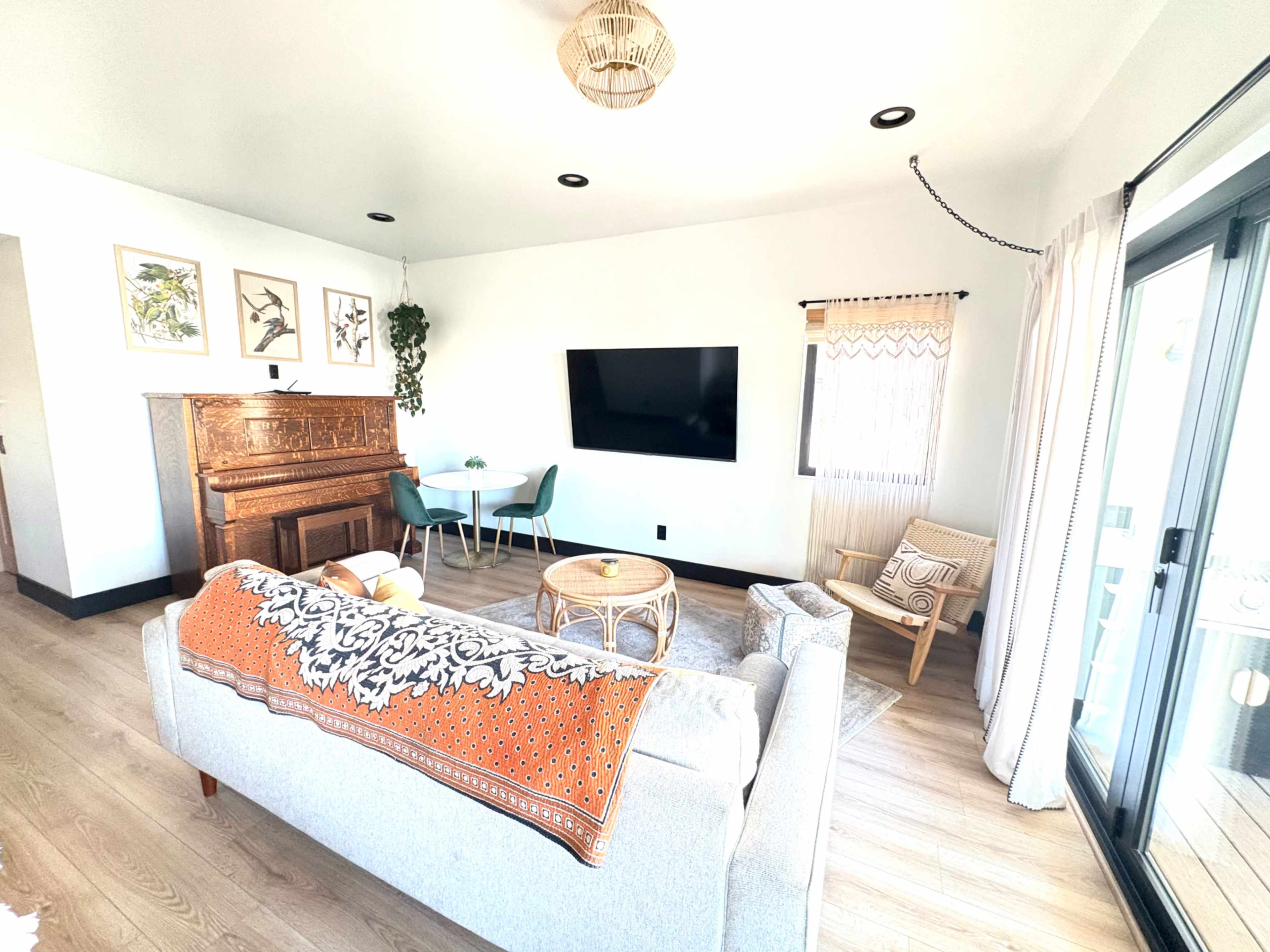 The living room features a gray sofa with an orange throw, a wooden piano, a large TV mounted on the wall, a small dining table with chairs, and natural light coming through glass doors.