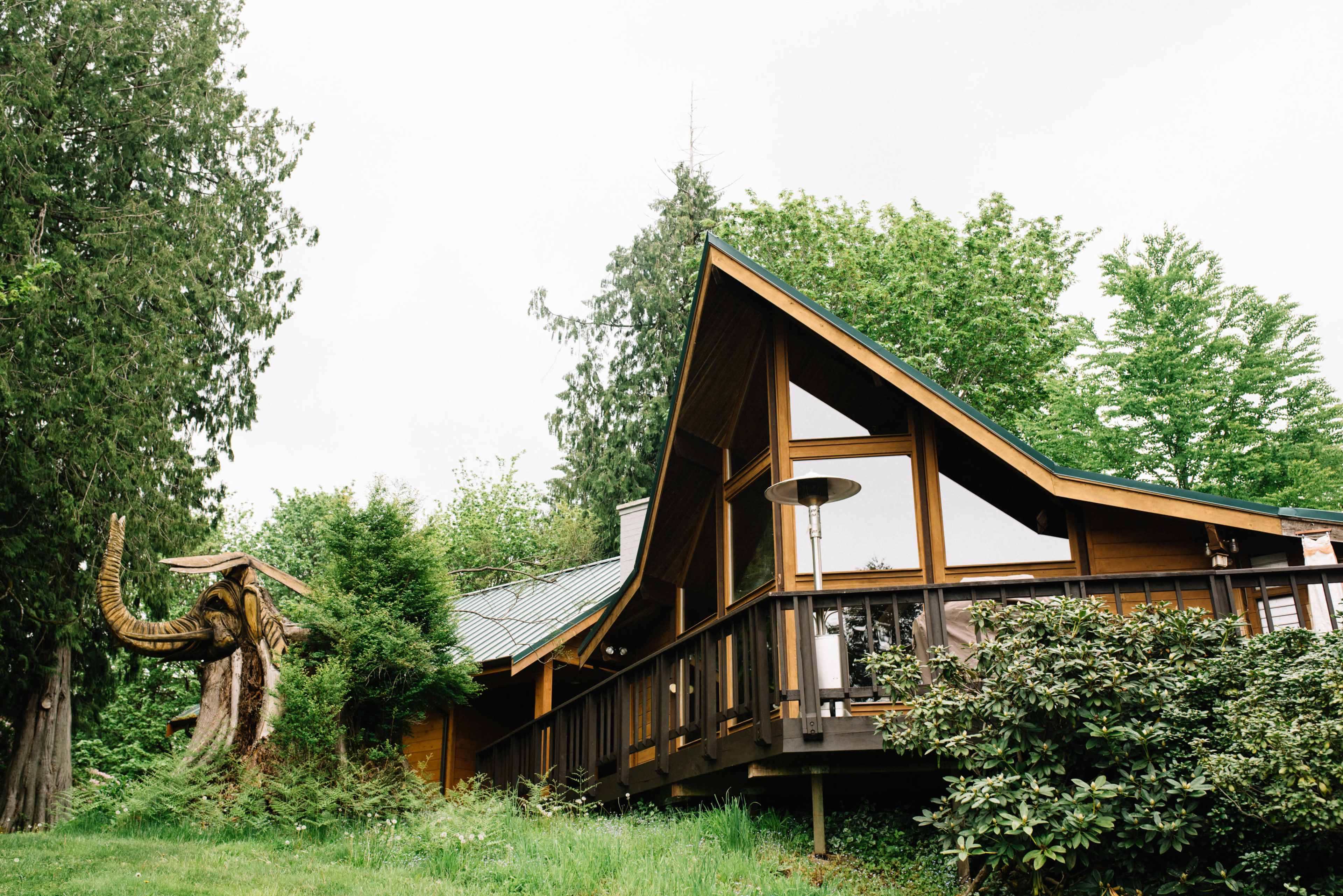 A wooden house with a distinctive sloped roof is surrounded by greenery and features a carved mammoth sculpture nearby.