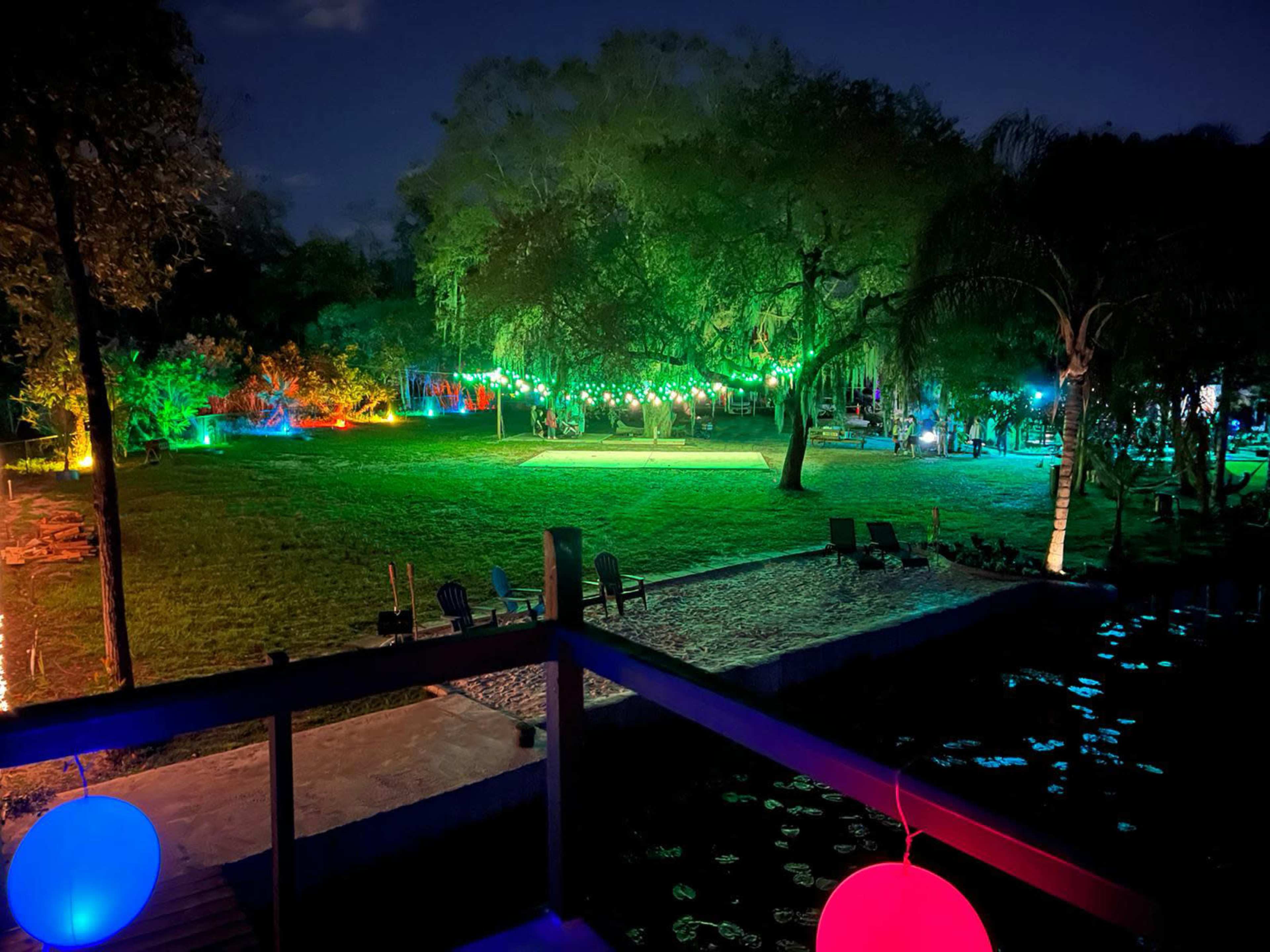 A night scene of a spacious outdoor area illuminated by colorful lights, with a grassy field and a water feature nearby.