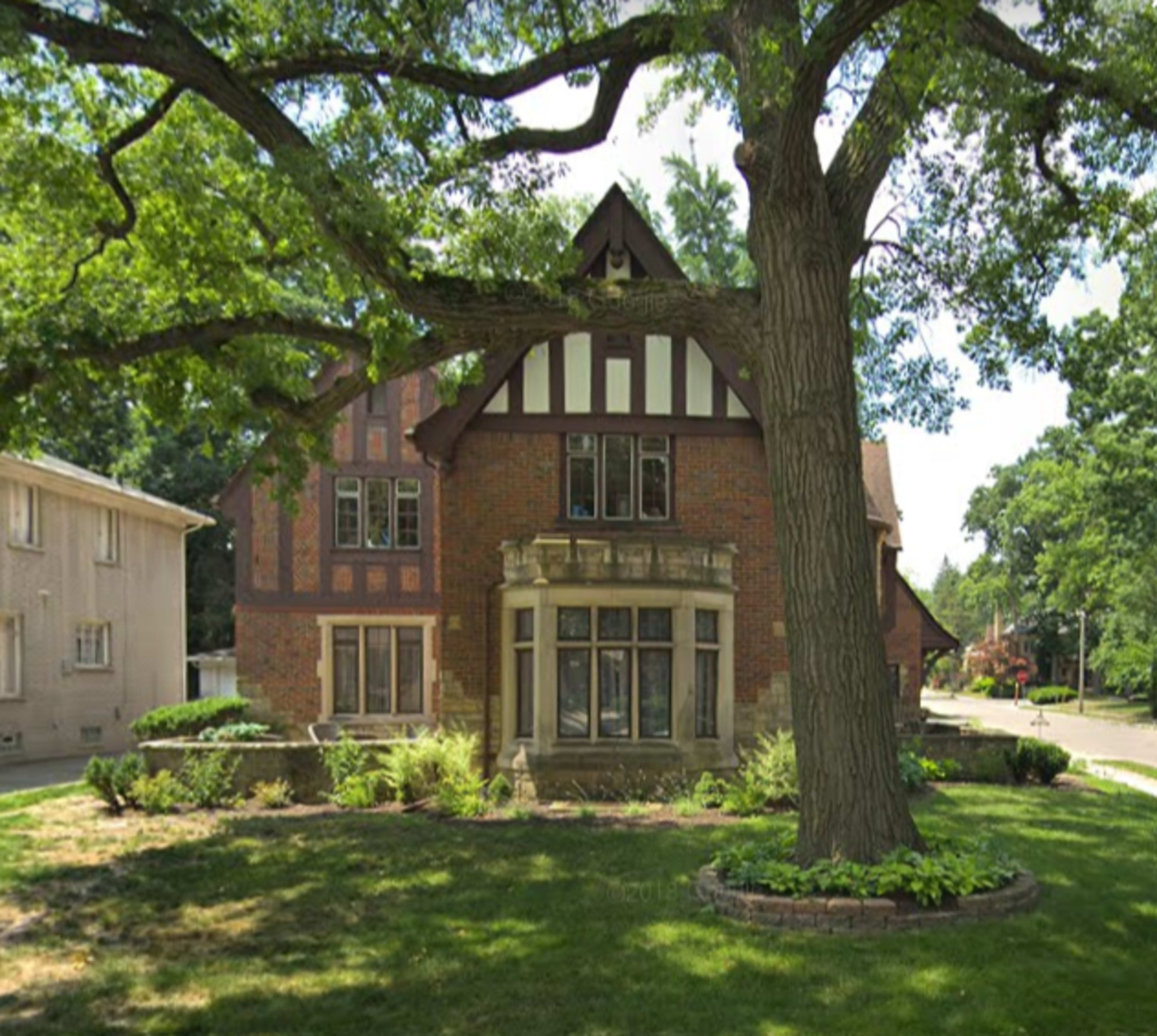 A large brick house with a steeply pitched roof and decorative timber accents is surrounded by a well-maintained lawn and mature trees.