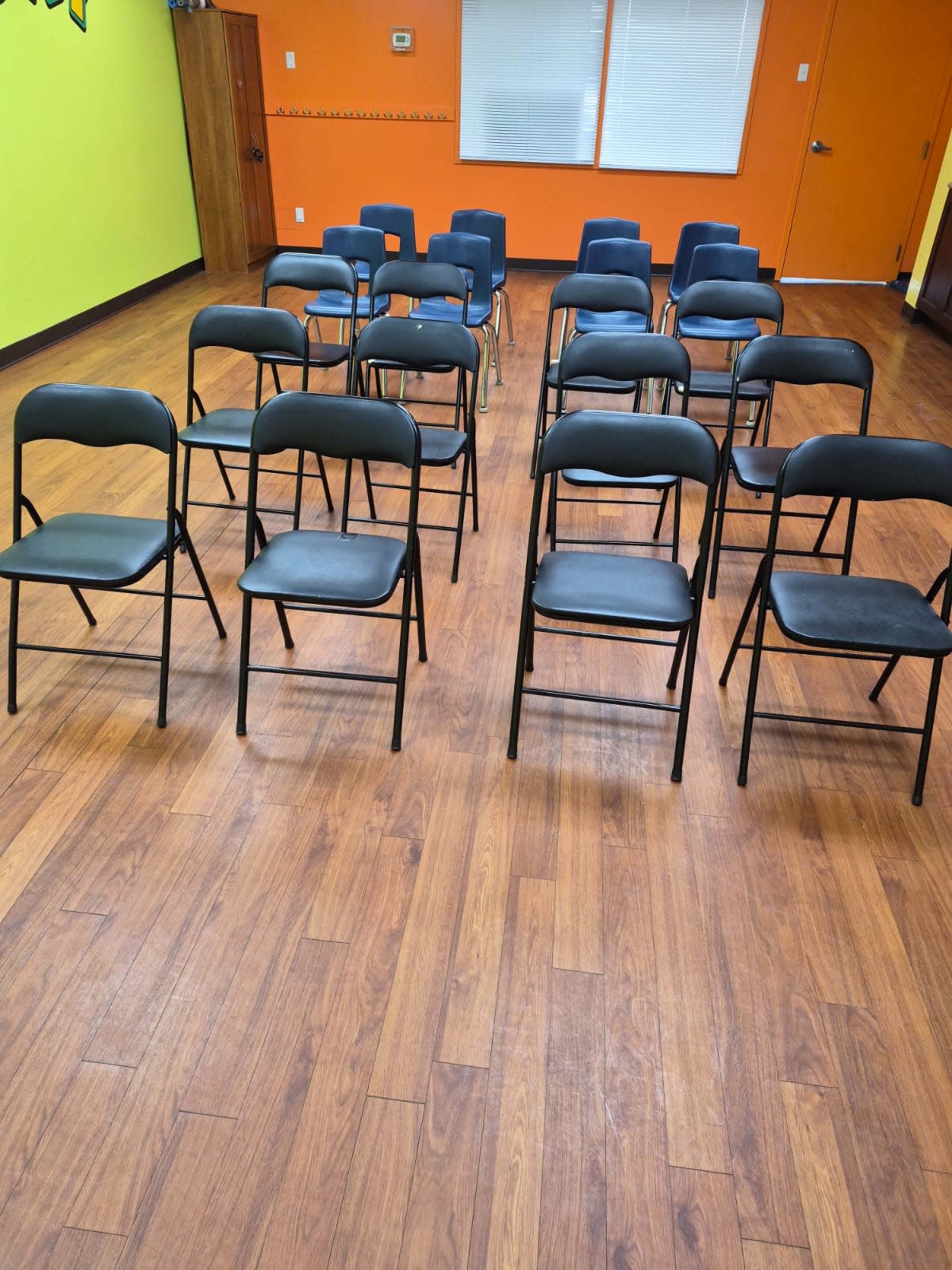 The image shows a room with wooden floors arranged in a grid of black folding chairs against brightly colored green and orange walls.
