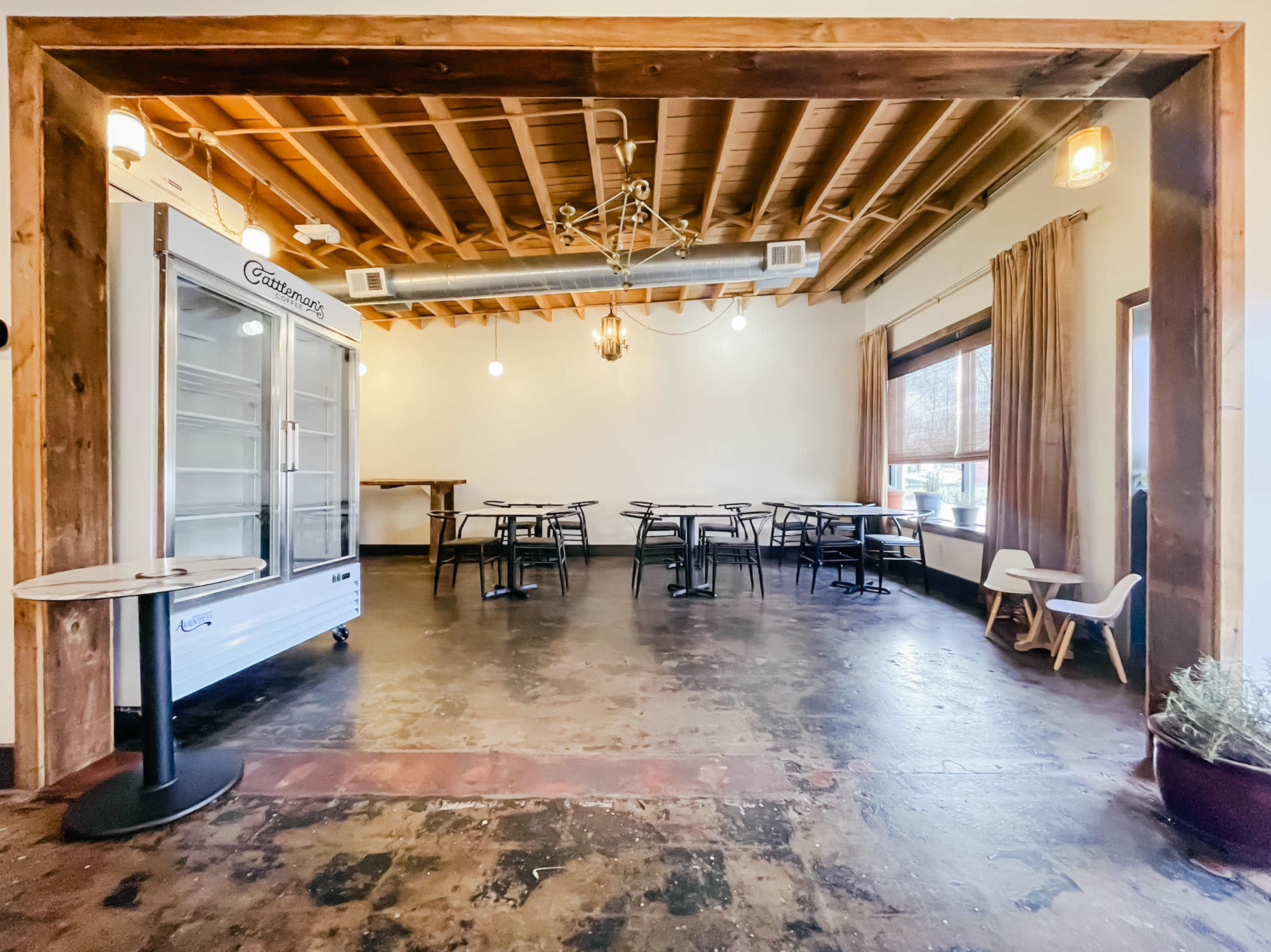 An empty, well-lit café space featuring tables and chairs arranged on a concrete floor, with a glass-front refrigerator and large windows.