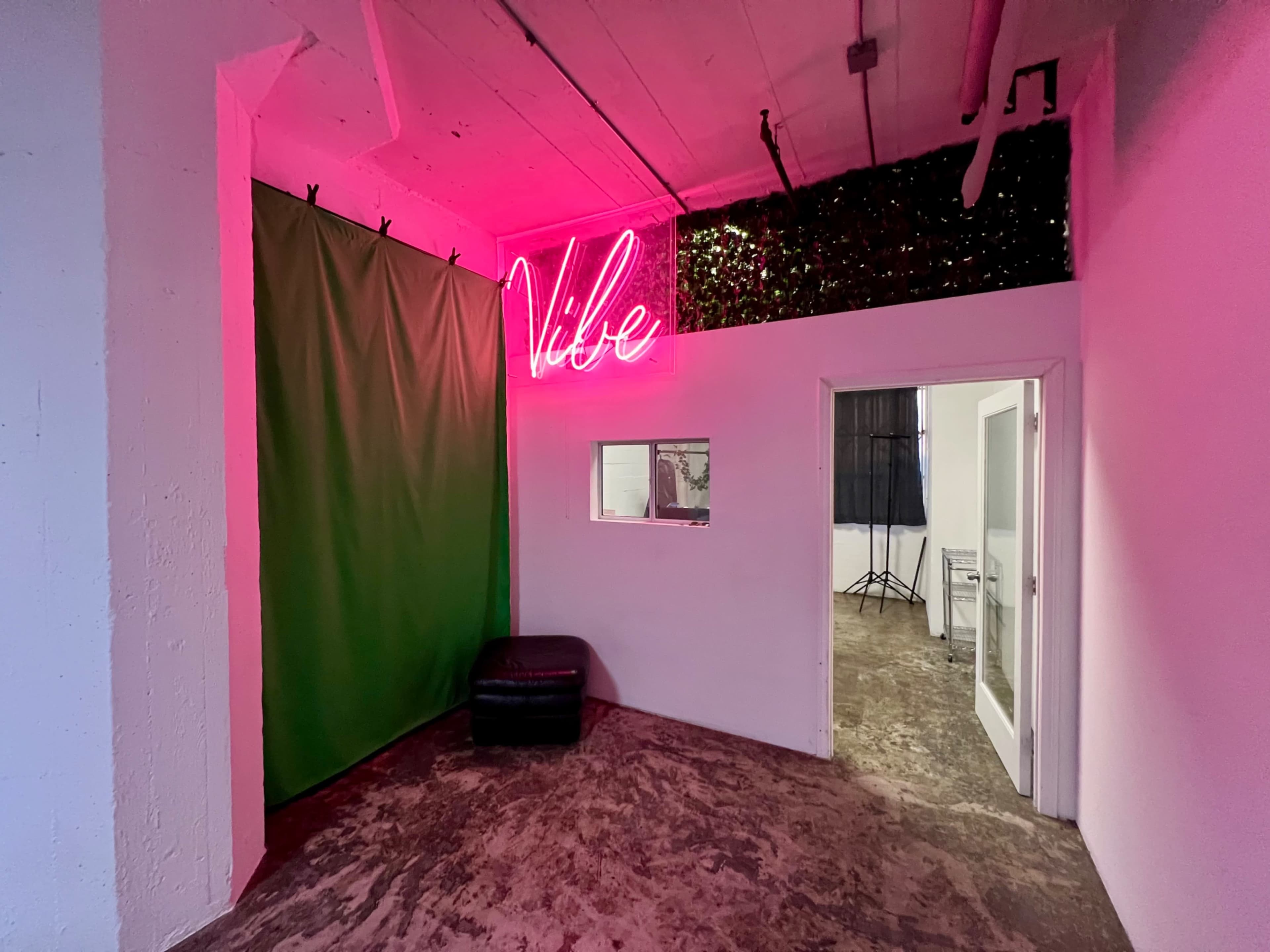 The image shows a corner of a studio space with a green backdrop, a neon pink "Vibe" sign, a small black chair, and a door leading to another area.