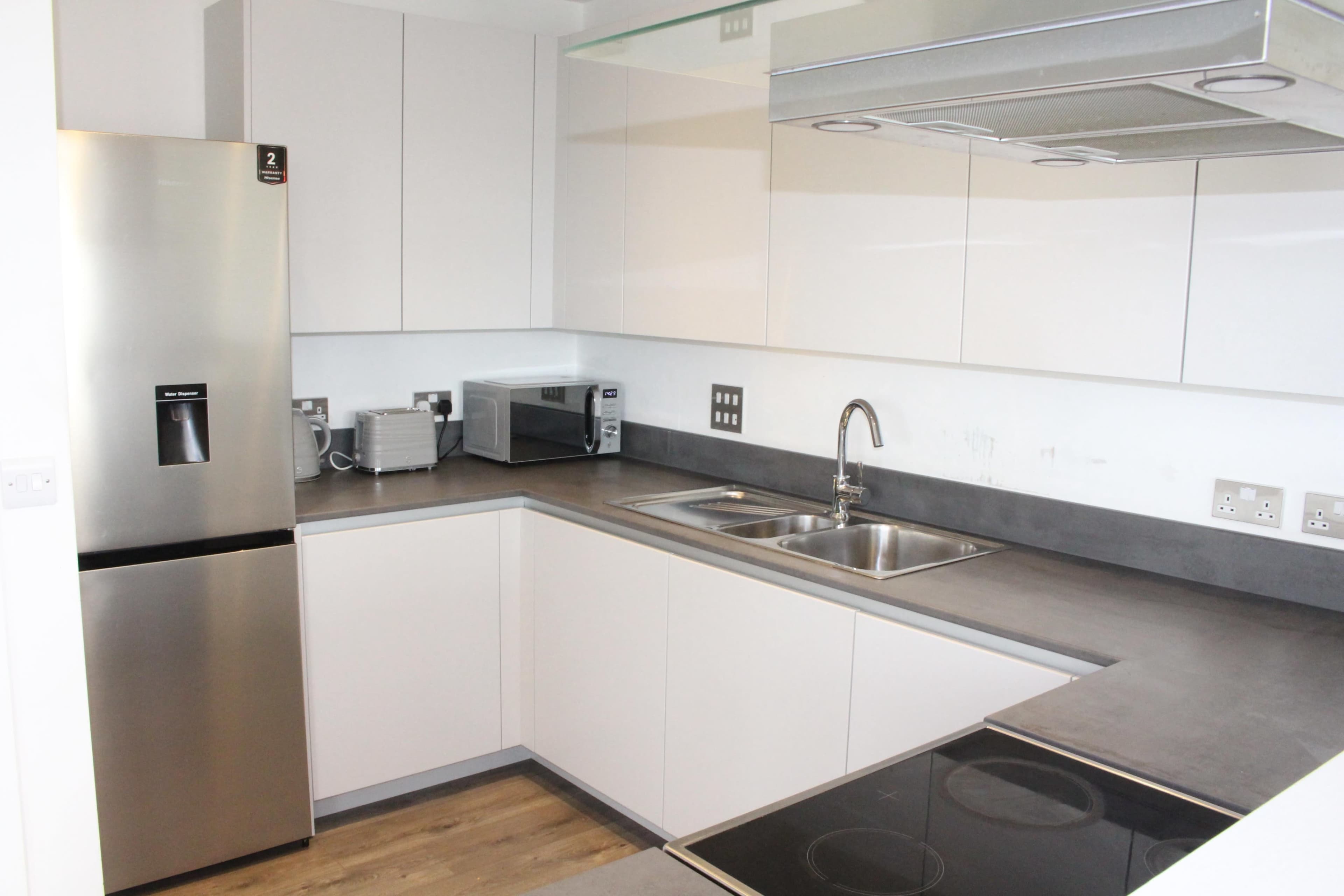 The image shows a modern kitchen with a stainless steel refrigerator, microwave, sink, and built-in stovetop, set against white cabinetry and gray countertops.