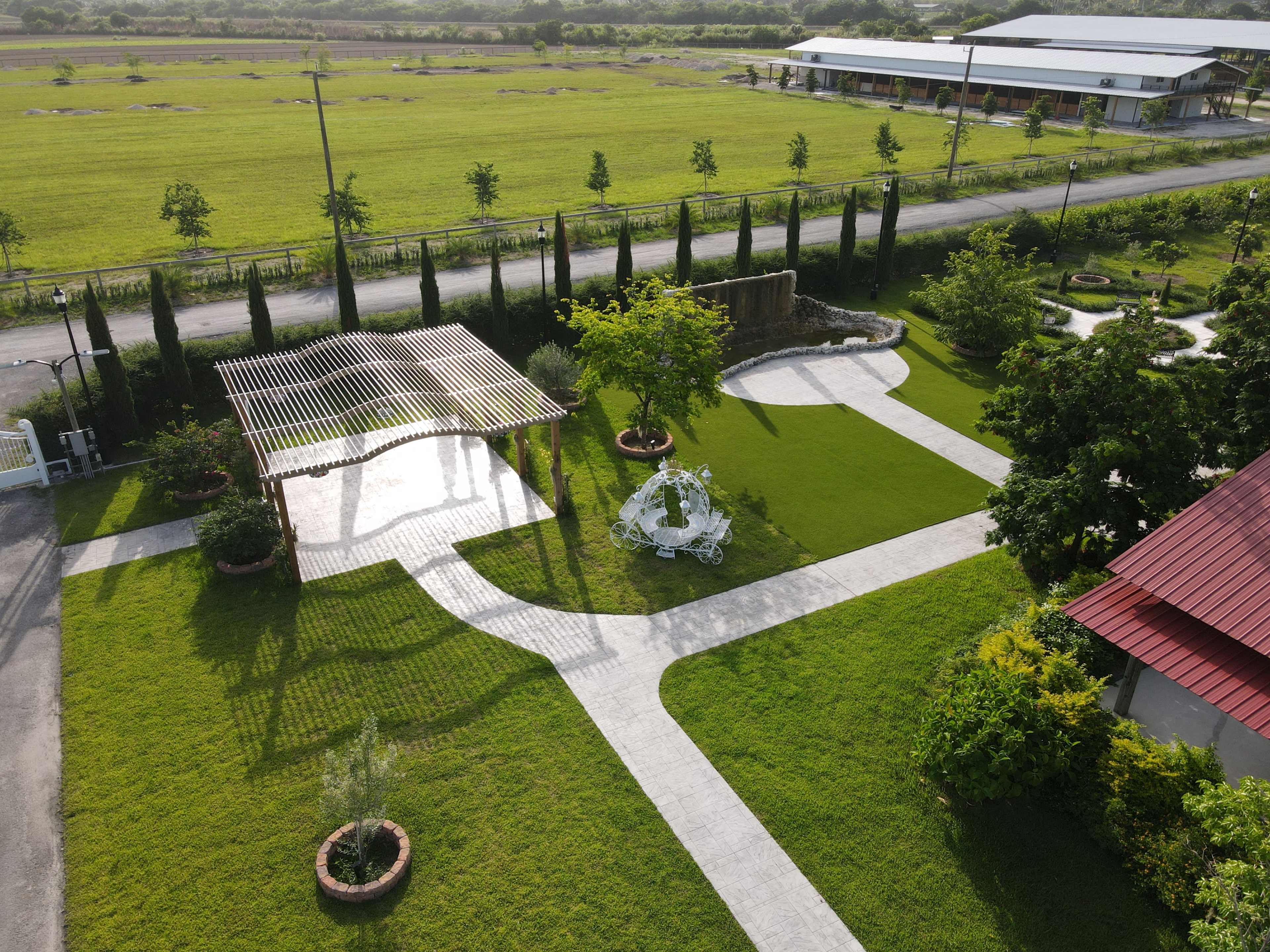 The image shows an aerial view of a landscaped garden with a paved pathway, greenery, a pergola, and a stone feature, surrounded by open fields and a building in the background.