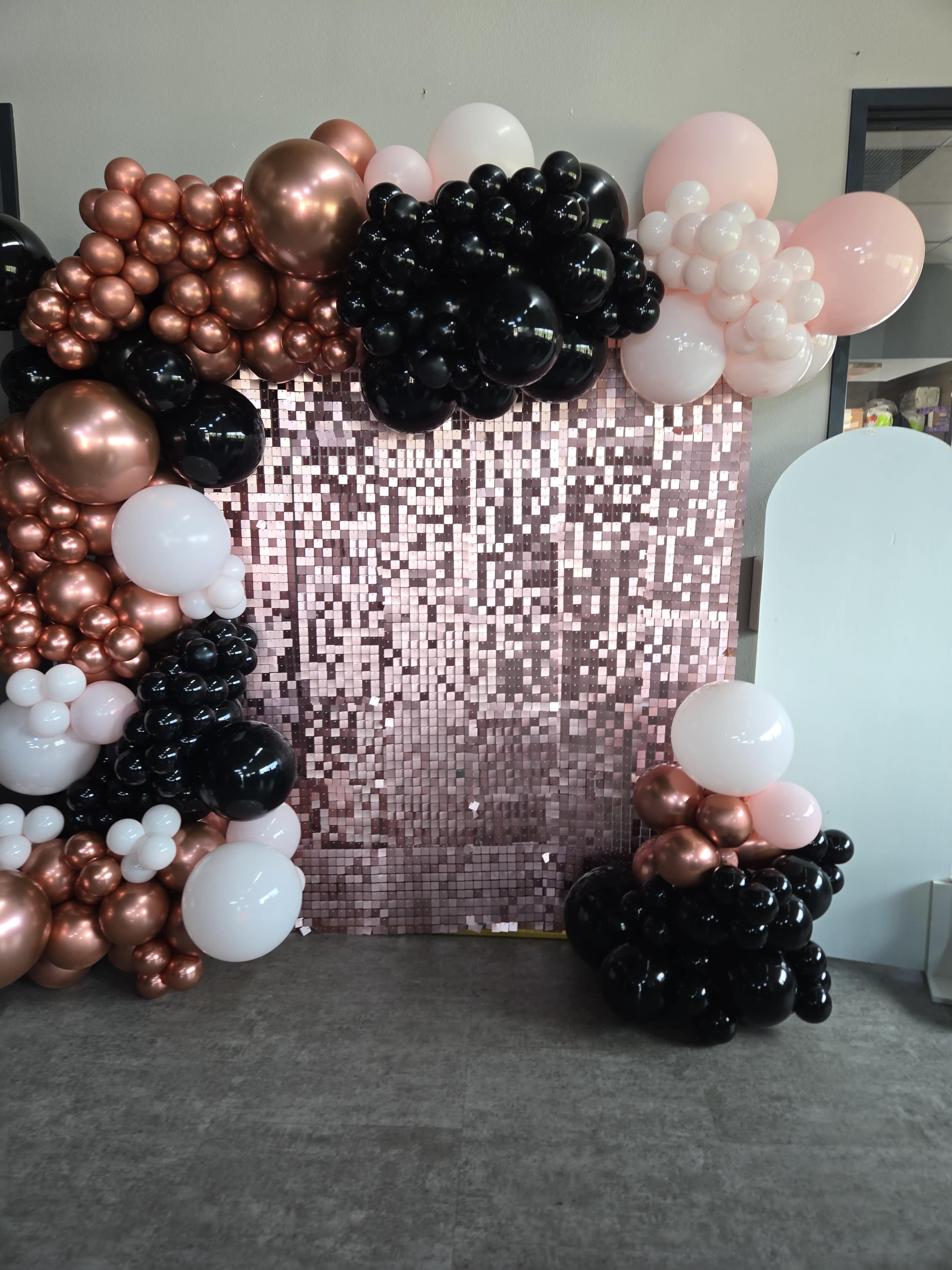 The image shows a party backdrop made of rose gold and black balloons arranged around a sparkling sequined curtain.