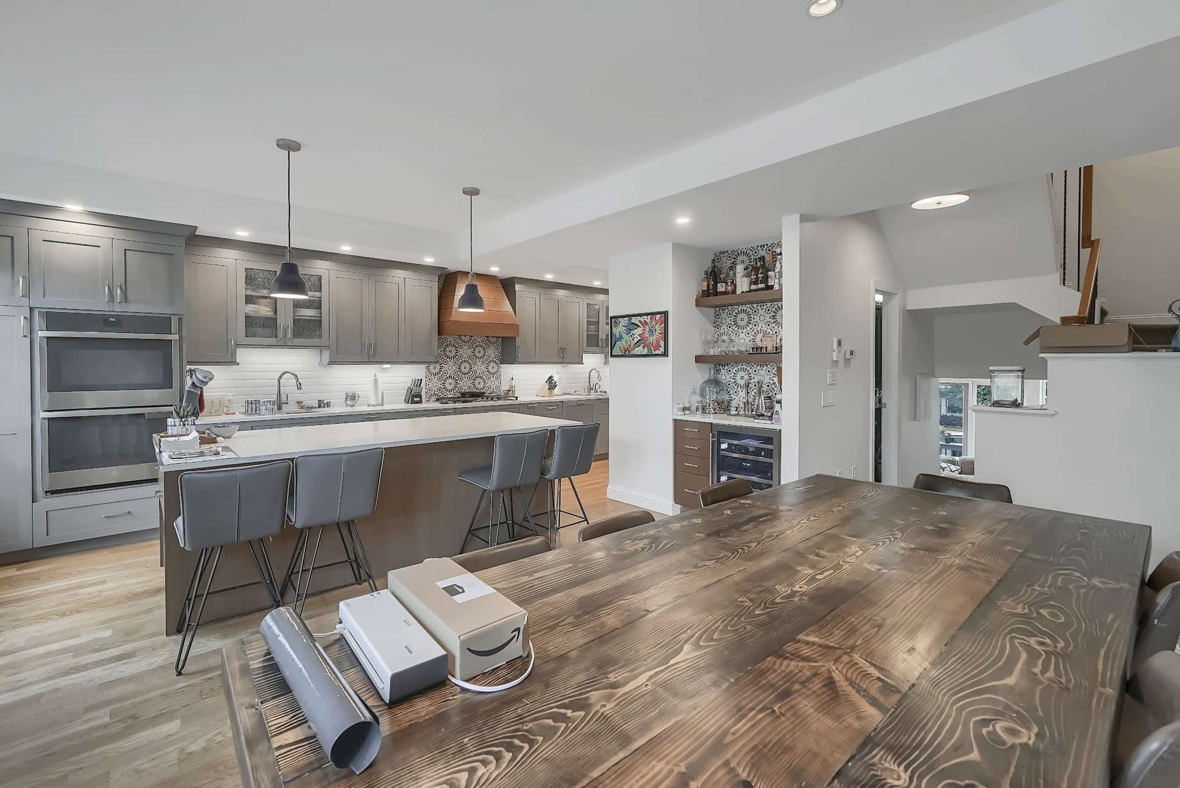 A modern kitchen features a large island with bar stools, wooden dining table, and stylish light fixtures.