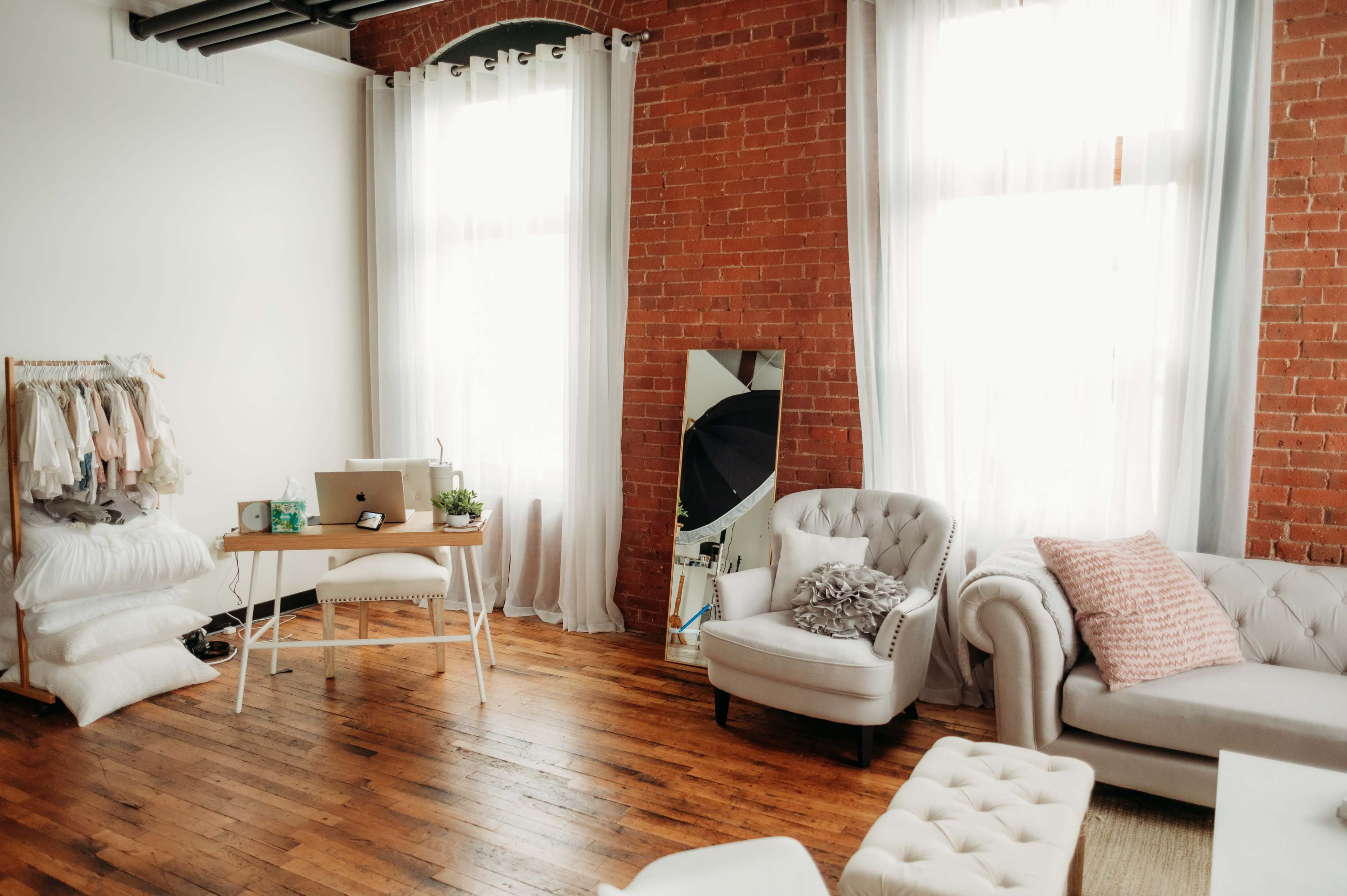 A cozy room features a wooden floor, large windows with white curtains, a desk with a computer, a mirror, and light-colored furniture.