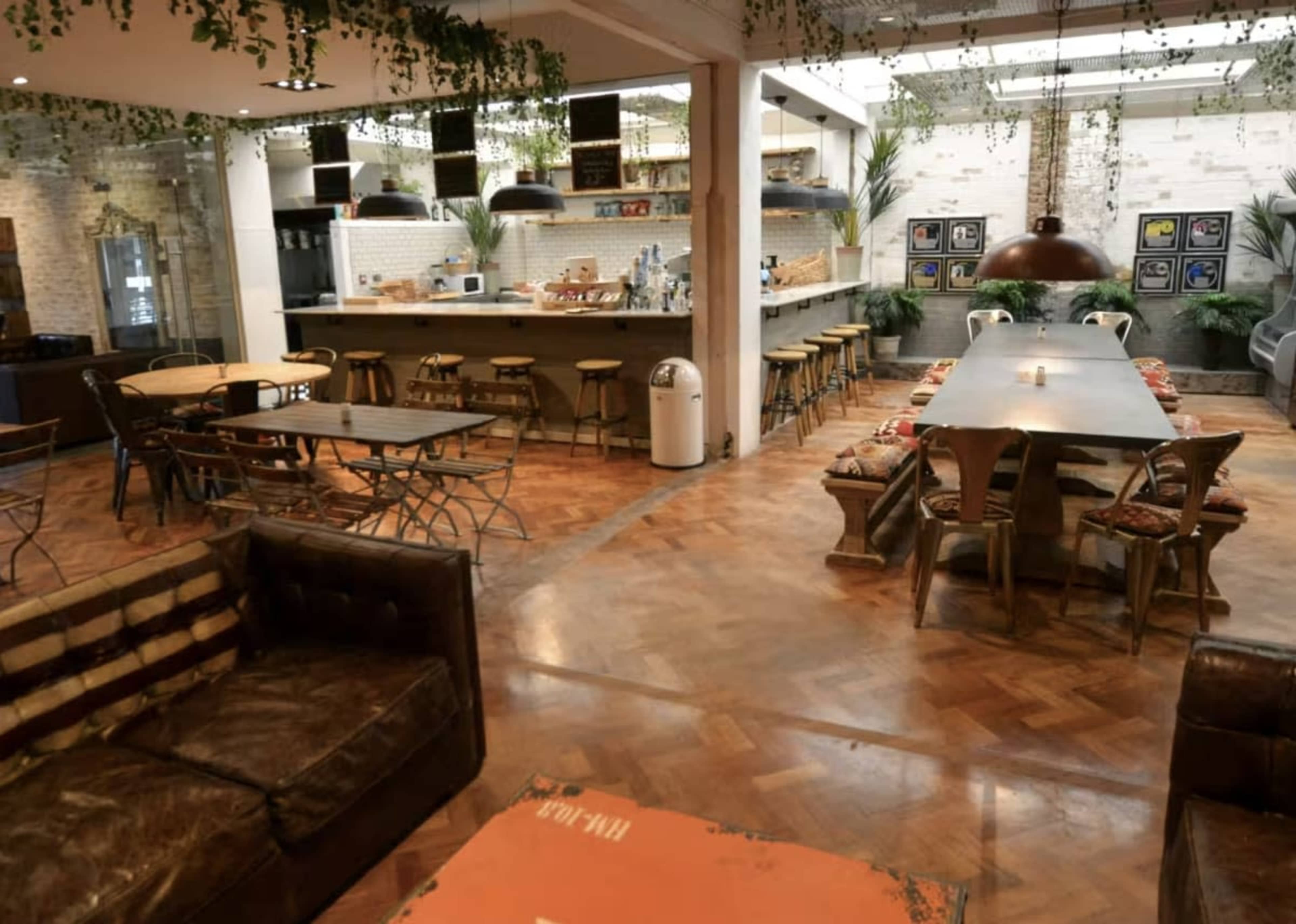 The image shows a spacious, modern interior of a café or communal dining area featuring wooden furniture, a large communal table, and a kitchen in the background.