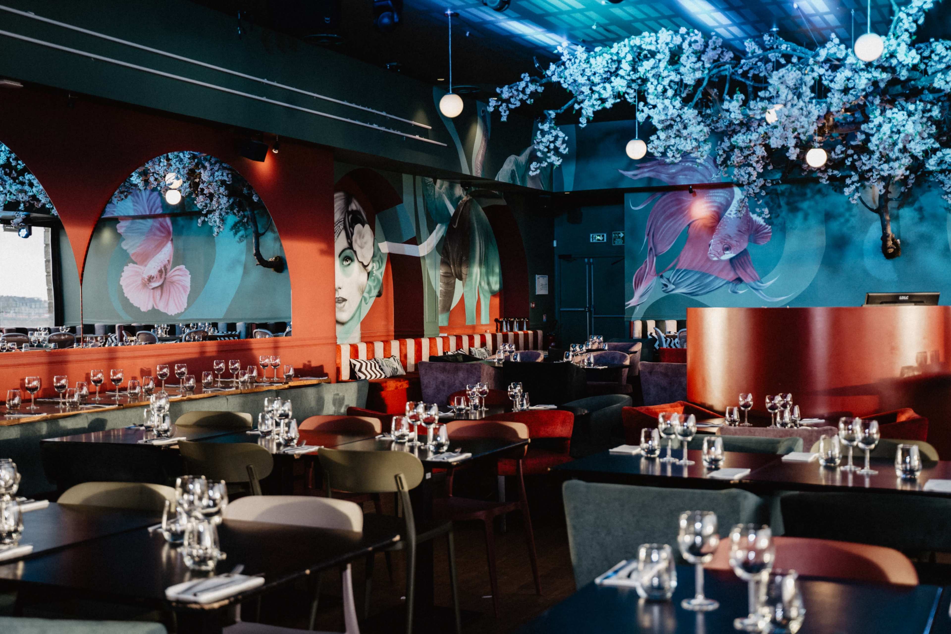 The image showcases a modern restaurant featuring vibrant murals, decorative floral elements, and elegantly set tables with glassware.