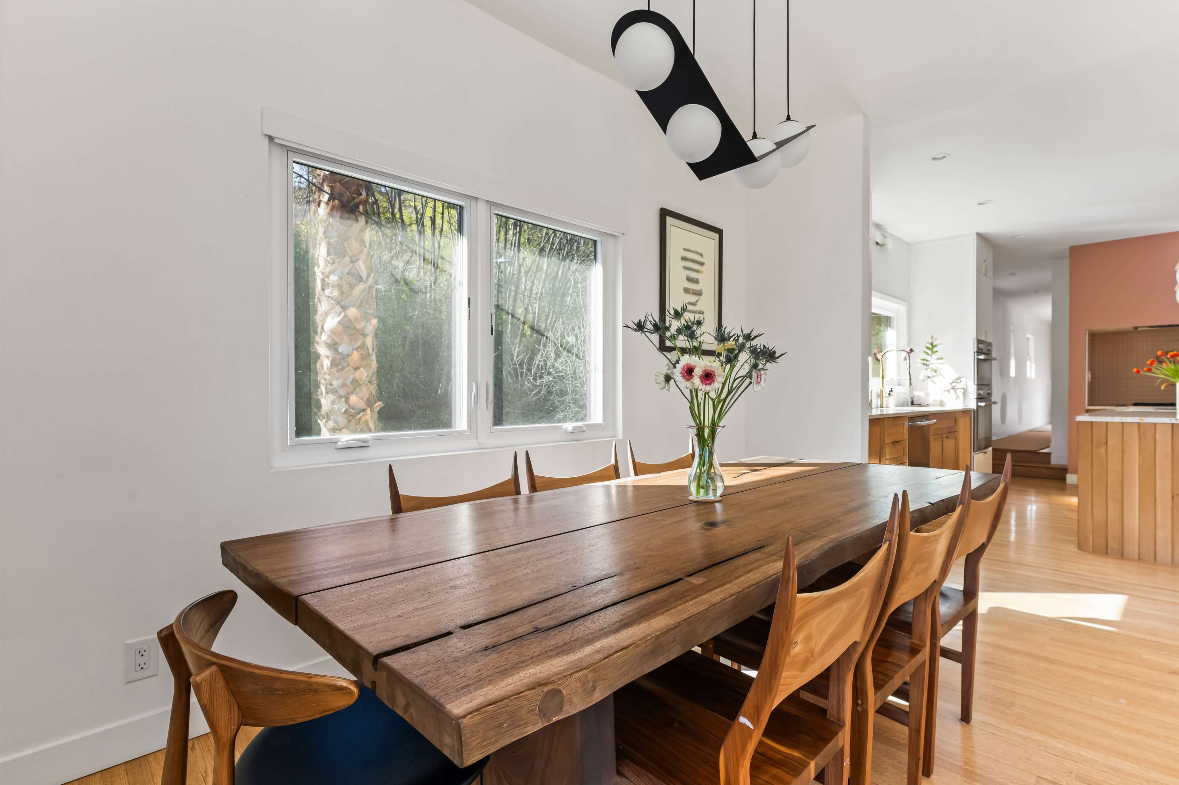 A wooden dining table with eight chairs sits in a brightly lit room, featuring large windows and a vase of flowers at the center.