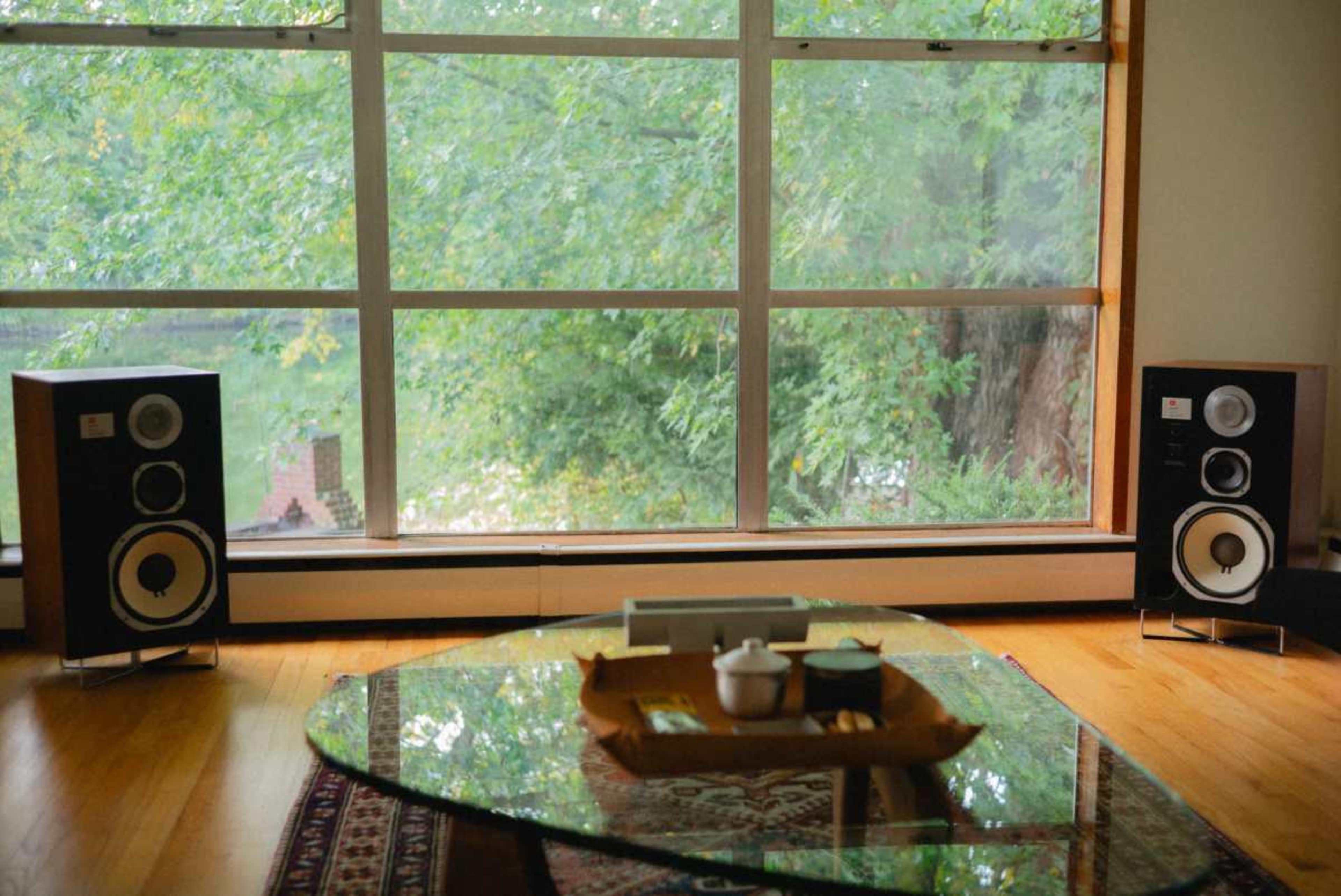 The image shows a living room featuring a large glass table, two speakers on either side, and a window overlooking greenery.