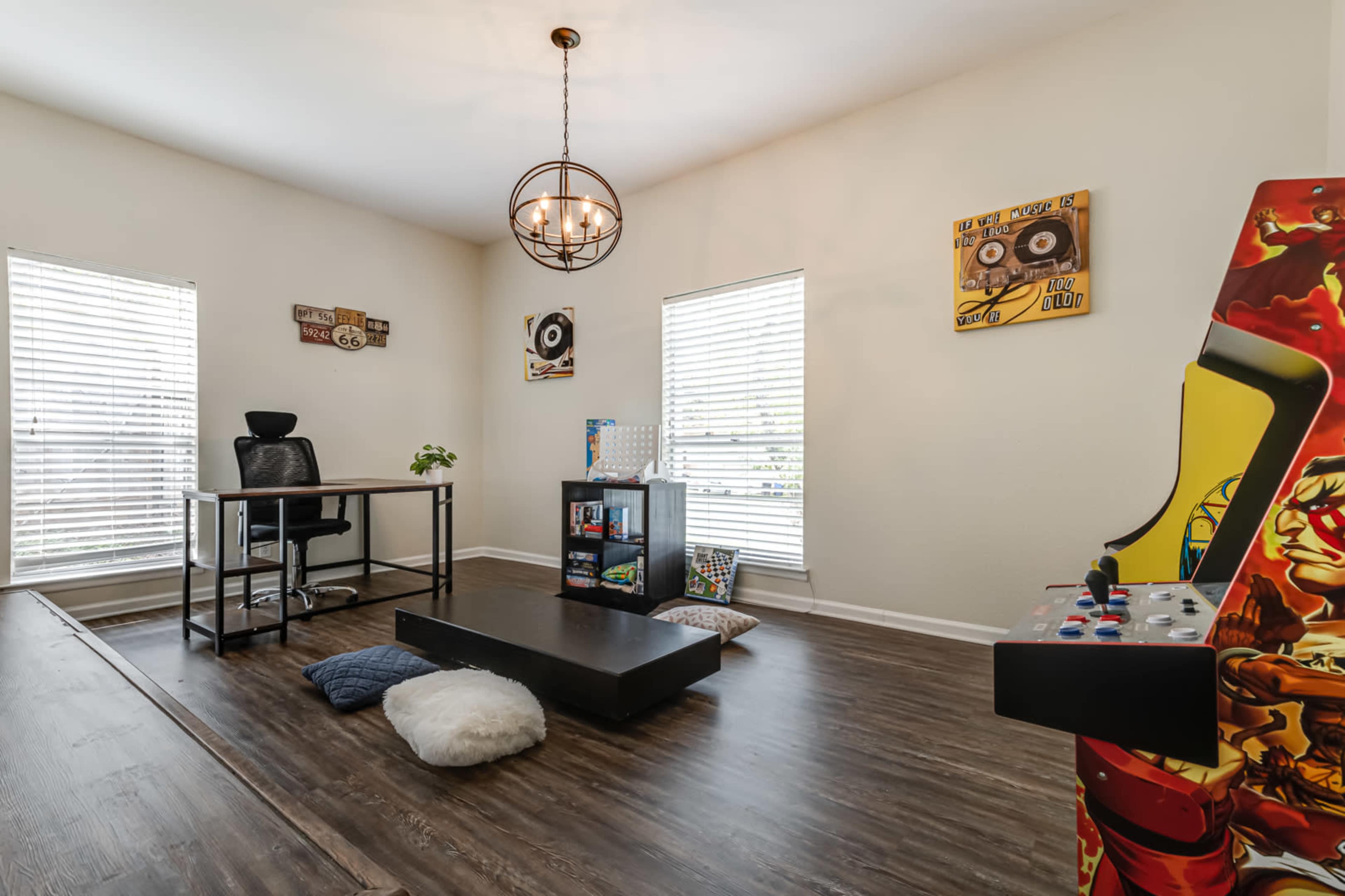 A minimalistic office space featuring a black desk and chair, a coffee table, a vintage arcade game machine, and two windows with blinds.
