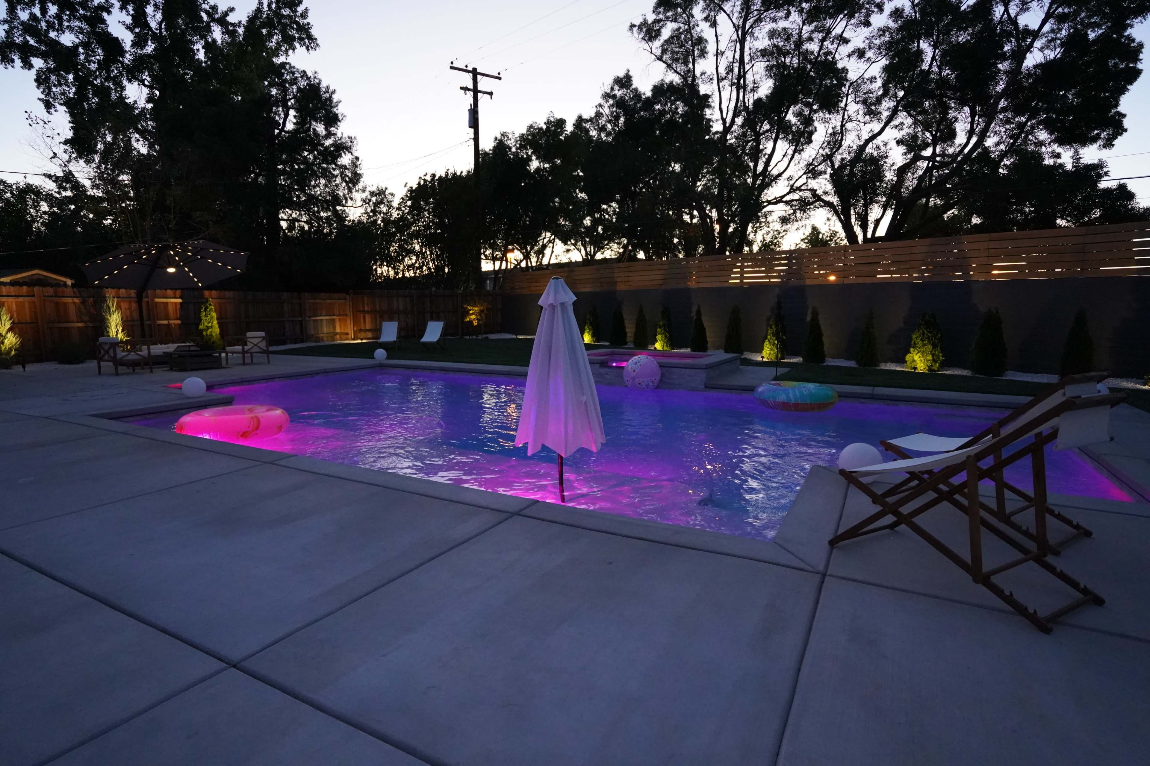 A swimming pool with illuminated water in shades of purple, surrounded by lounge chairs and a shaded area at dusk.