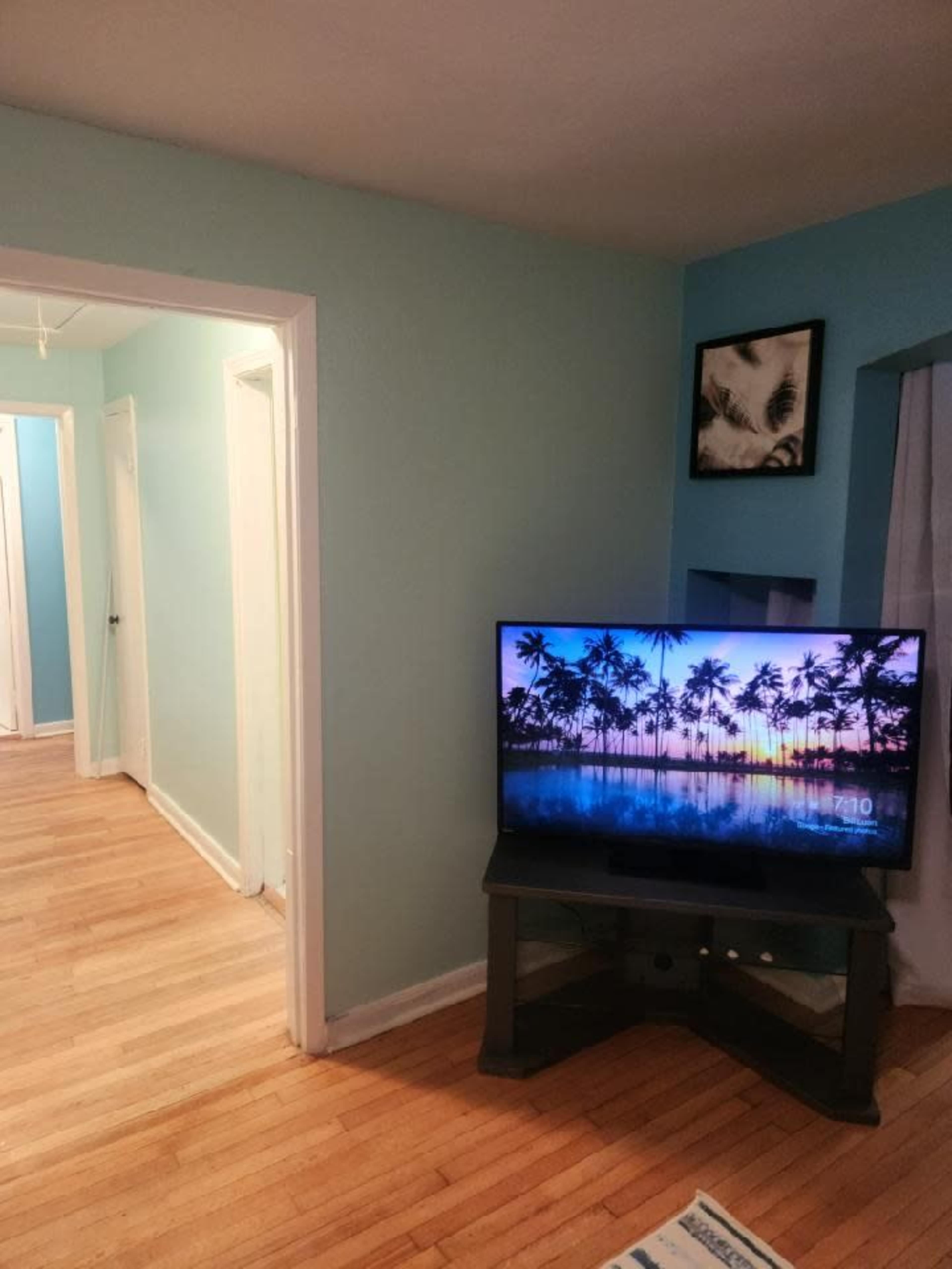The image shows a living room with a television displaying a tropical sunset scene and a view of a hallway with a light blue wall and wooden flooring.