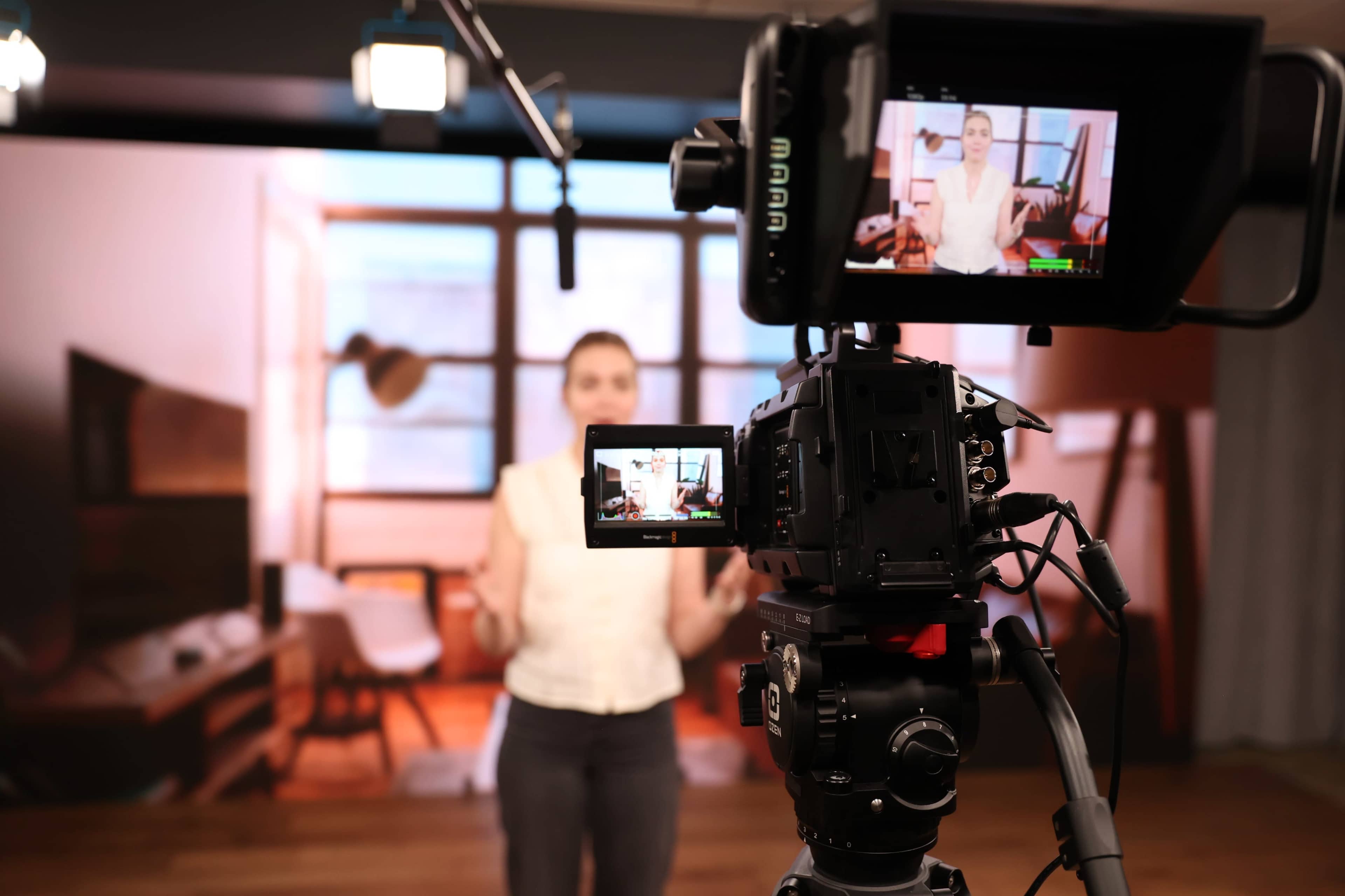 A camera is focused on a speaker in a studio setting with a backdrop of a well-furnished room.
