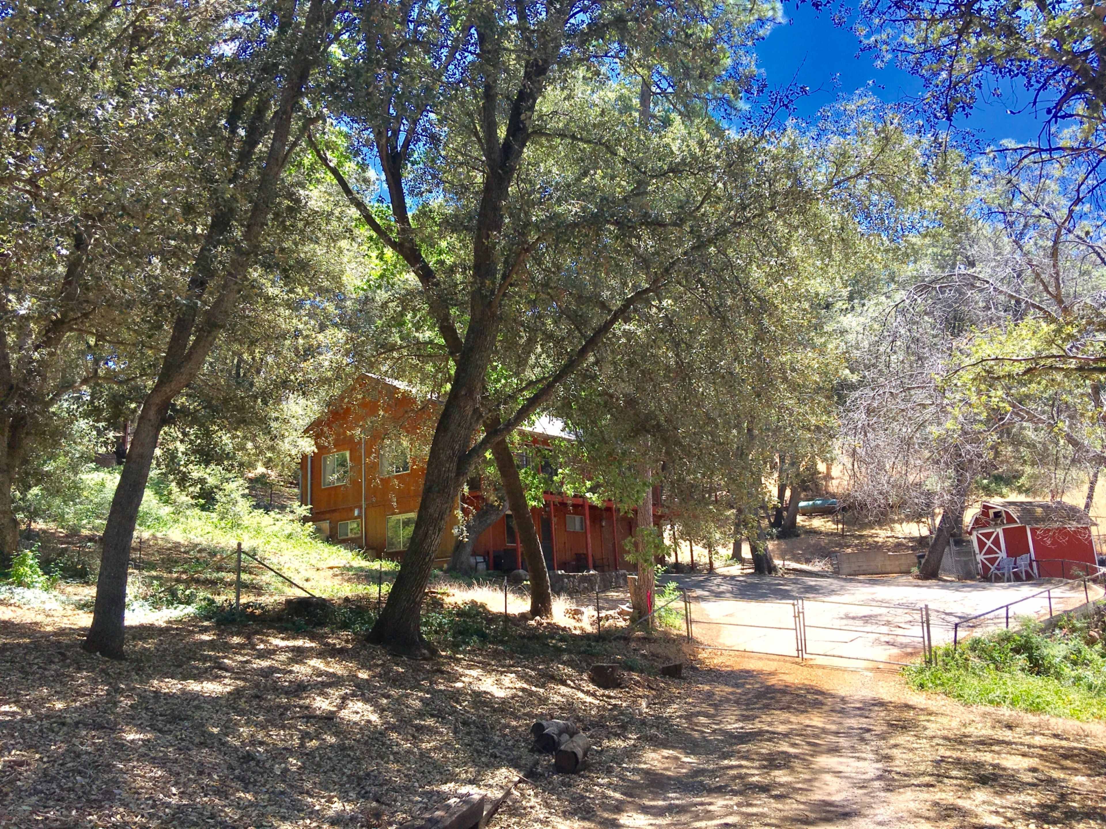 A wooden orange house sits among large trees, with a red barn visible in the background and a gravel path leading to a gated entrance.