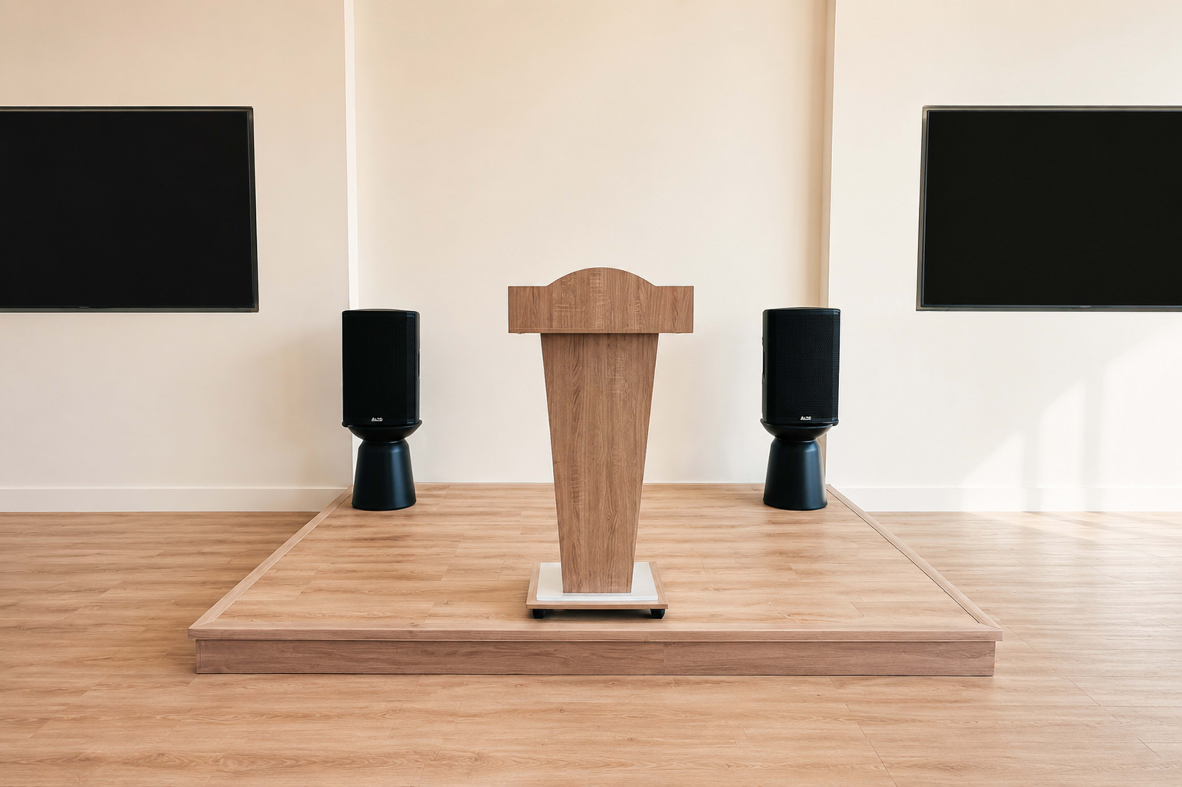 A wooden podium stands on a raised platform between two large speakers in a minimalist room with white walls.