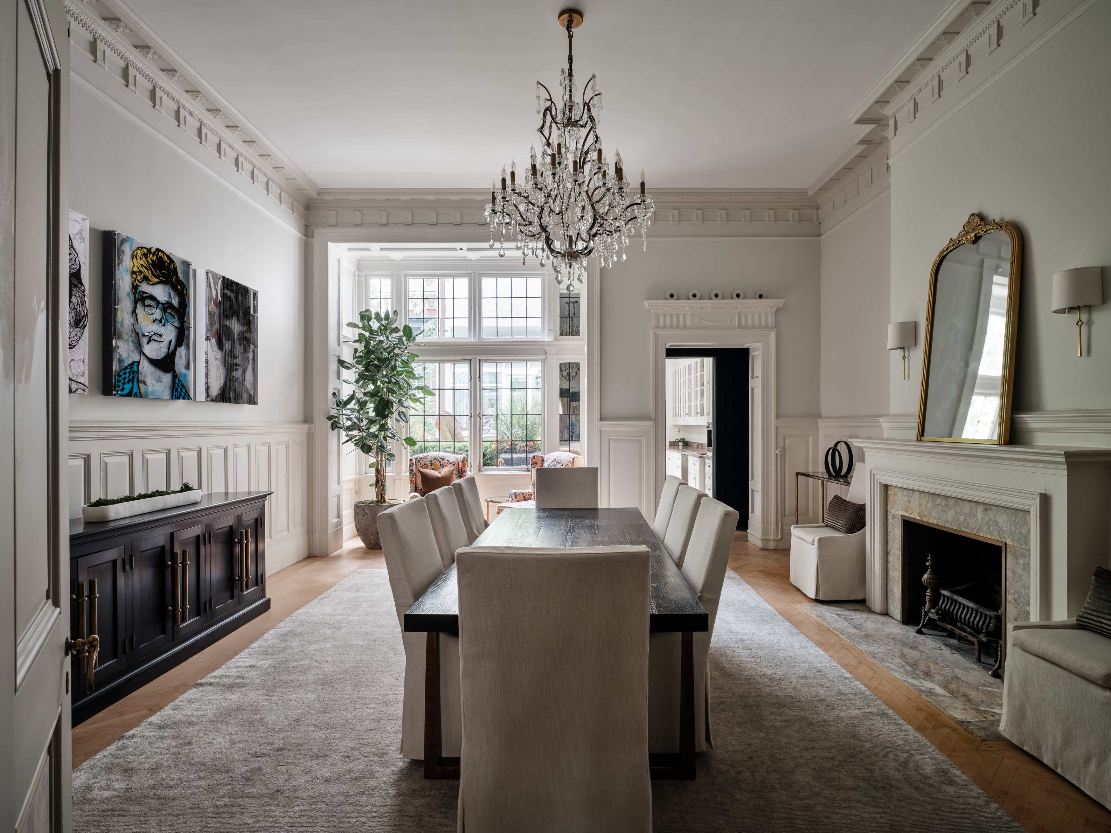 A spacious dining room features a large table surrounded by white upholstered chairs, illuminated by a crystal chandelier, and has large windows with views of a cozy sitting area.
