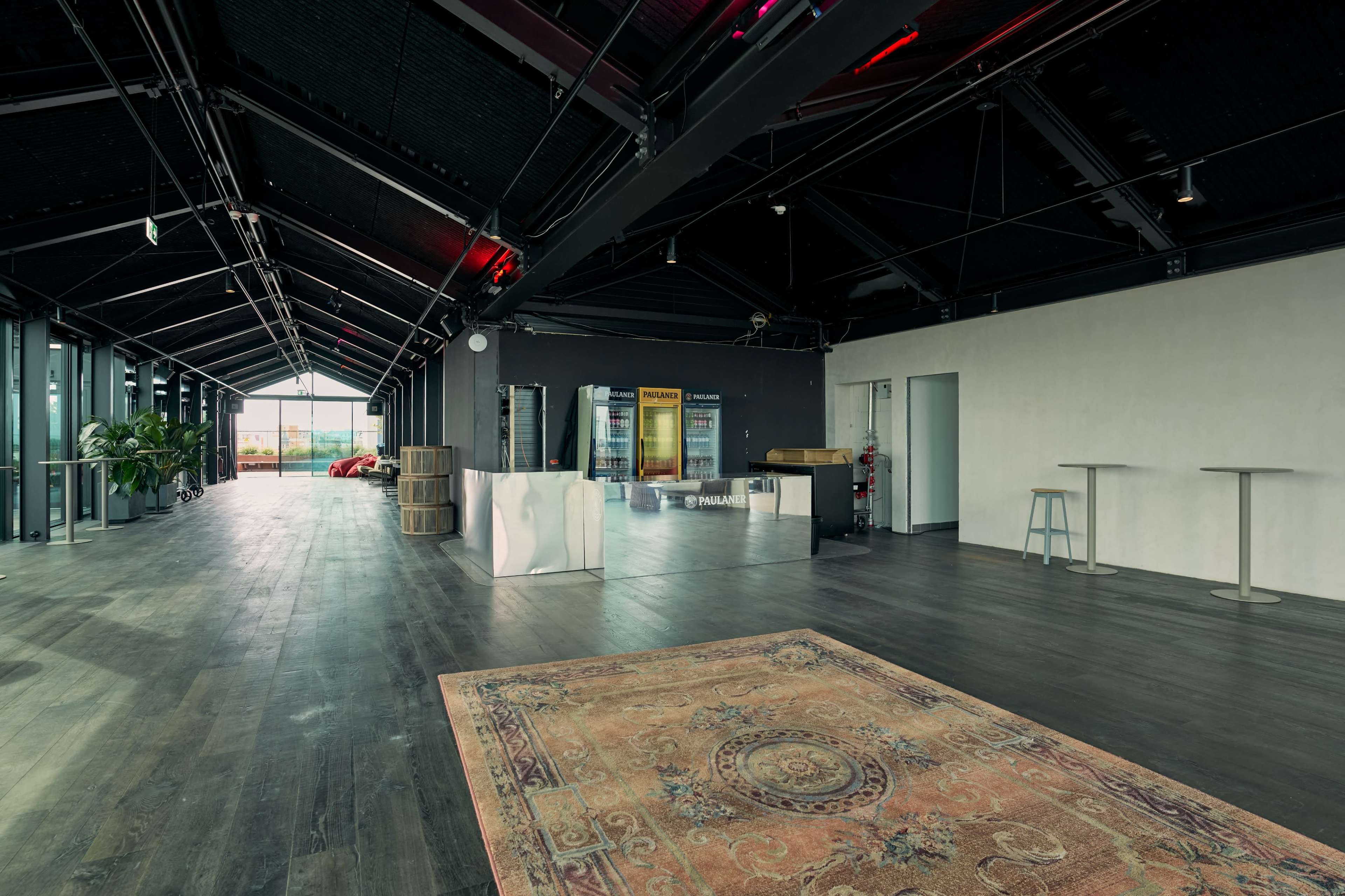 The image shows a spacious, modern interior with large windows, a reception area featuring a mirrored desk, and minimal furnishings including a few tables and a decorative rug on the floor.