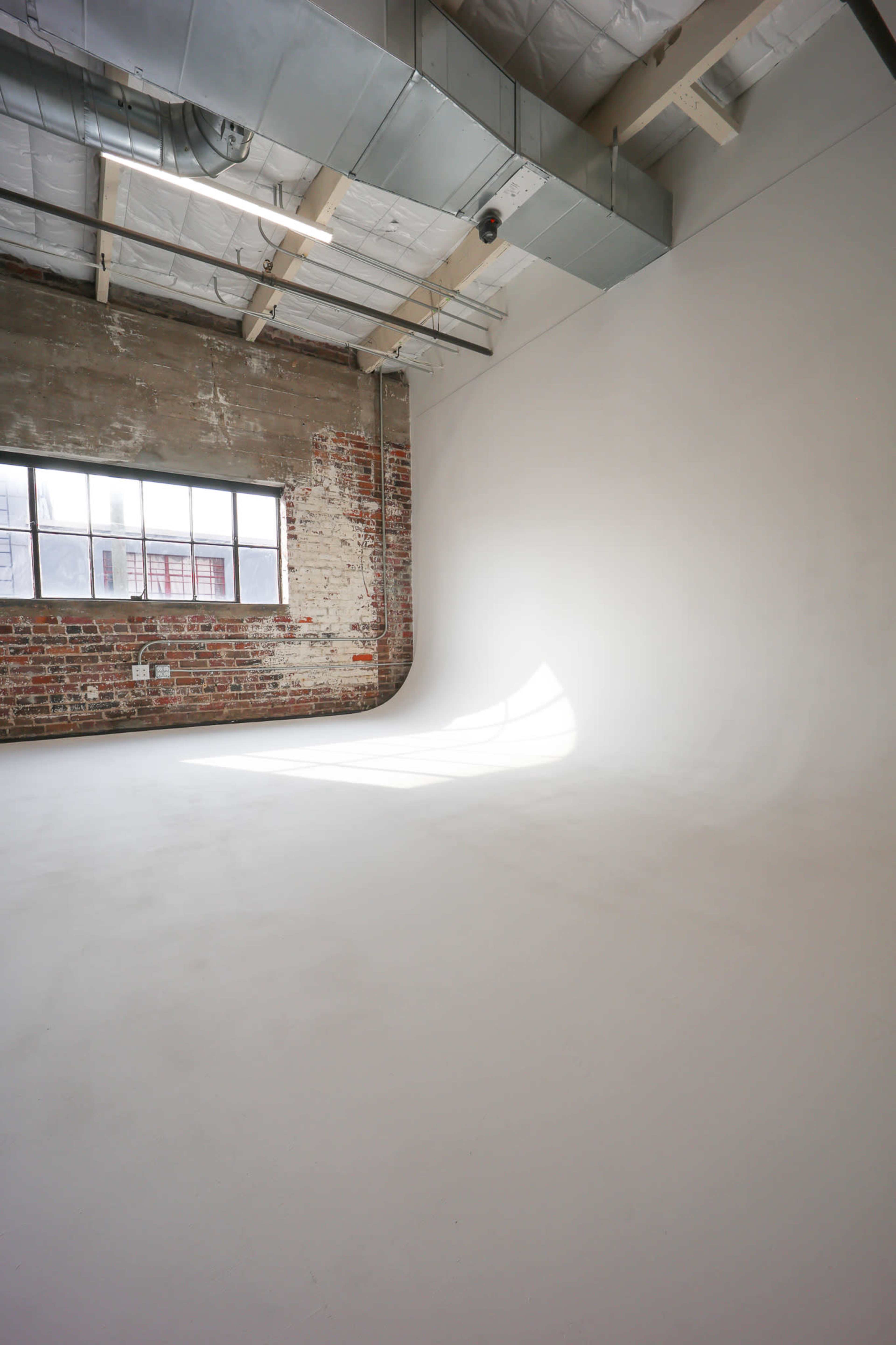 The image shows an empty studio space with a smooth white floor, exposed brick walls, and a large window letting in natural light.