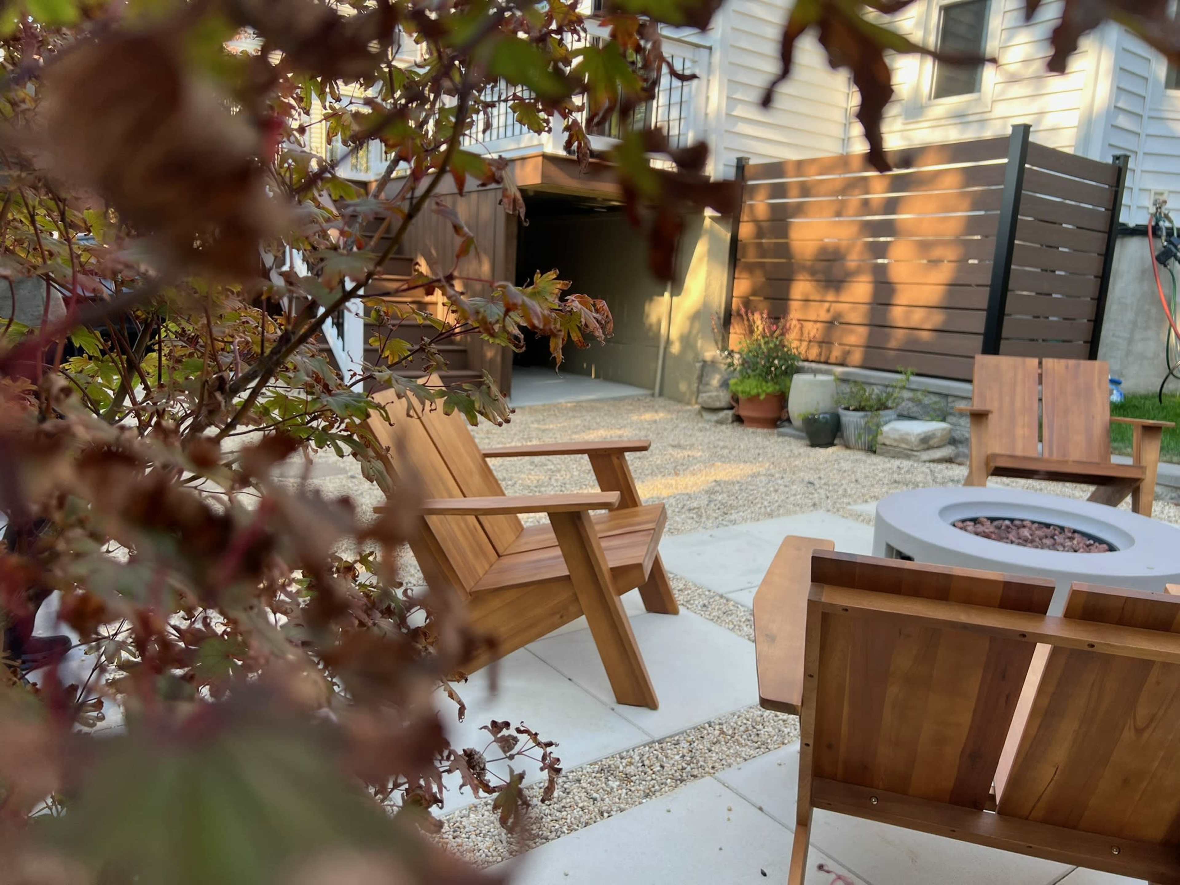 The image shows a cozy outdoor seating area featuring wooden chairs around a fire pit, with a backdrop of a garage and a landscaped garden.