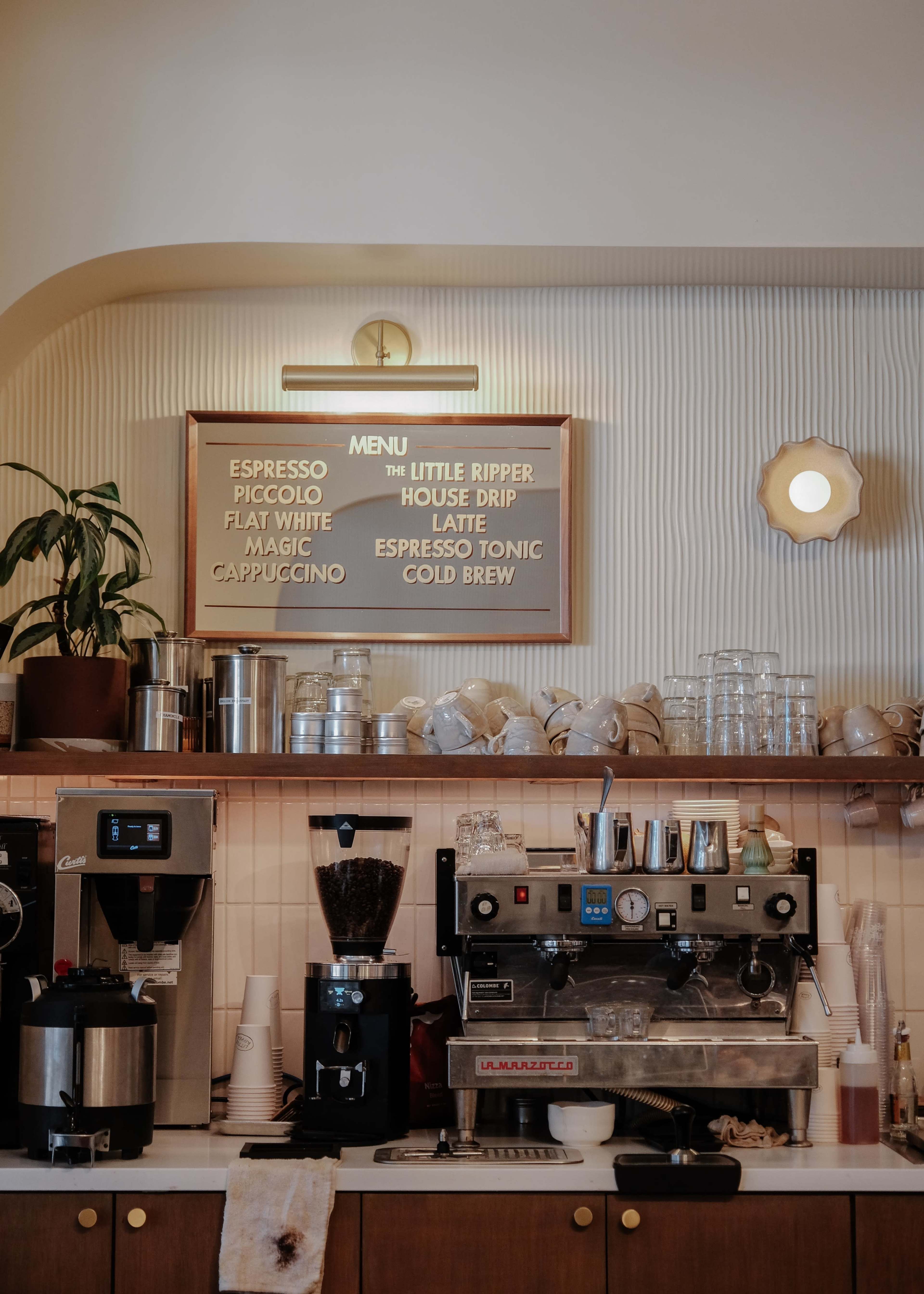 A coffee shop counter with a menu board displaying various coffee drinks above espresso machines and glassware.